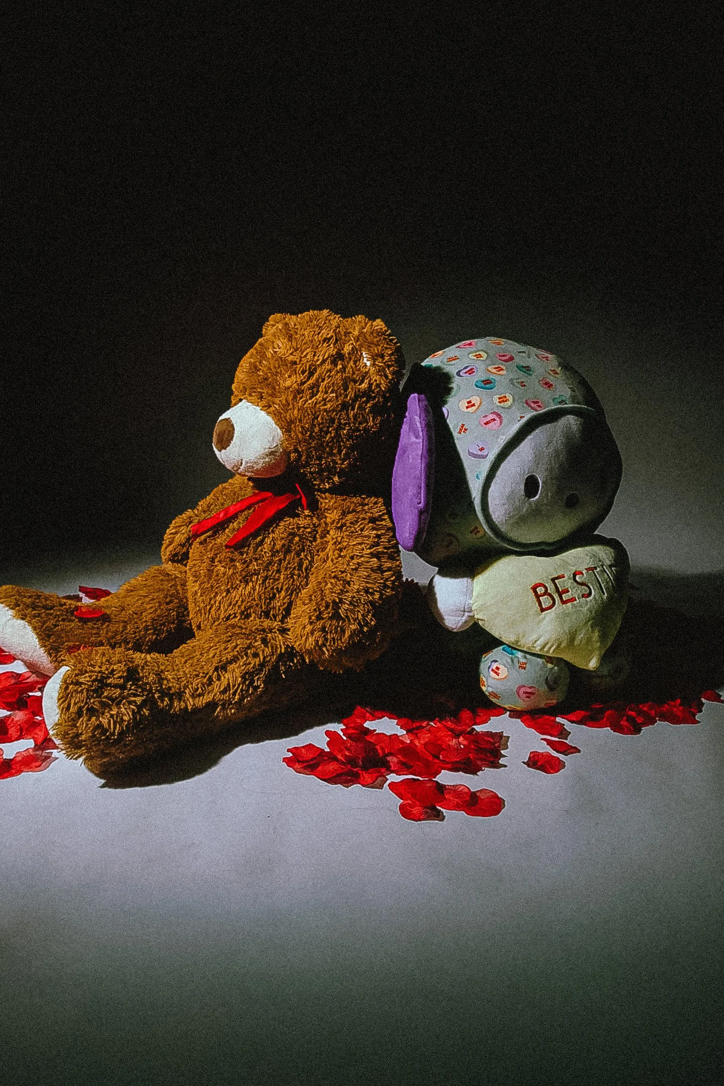 A large stuffed teddy bear with a red ribbon around its neck sits on a surface. Next to it is a balloon shaped like a dog, with colorful patterns and the word "BEST" printed on it. Red rose petals are scattered around them, against a dark background.