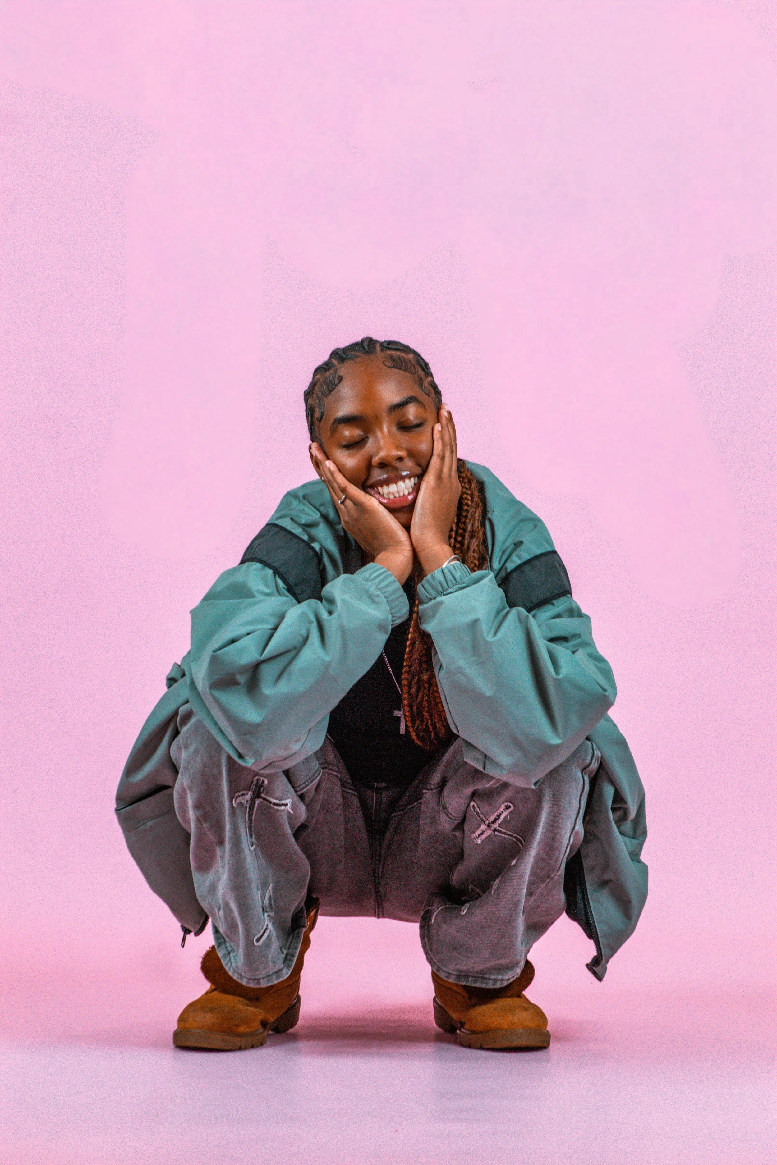 A smiling young woman with braided hair wearing a teal and black jacket, gray pants, and tan boots, squatting against a pink background.