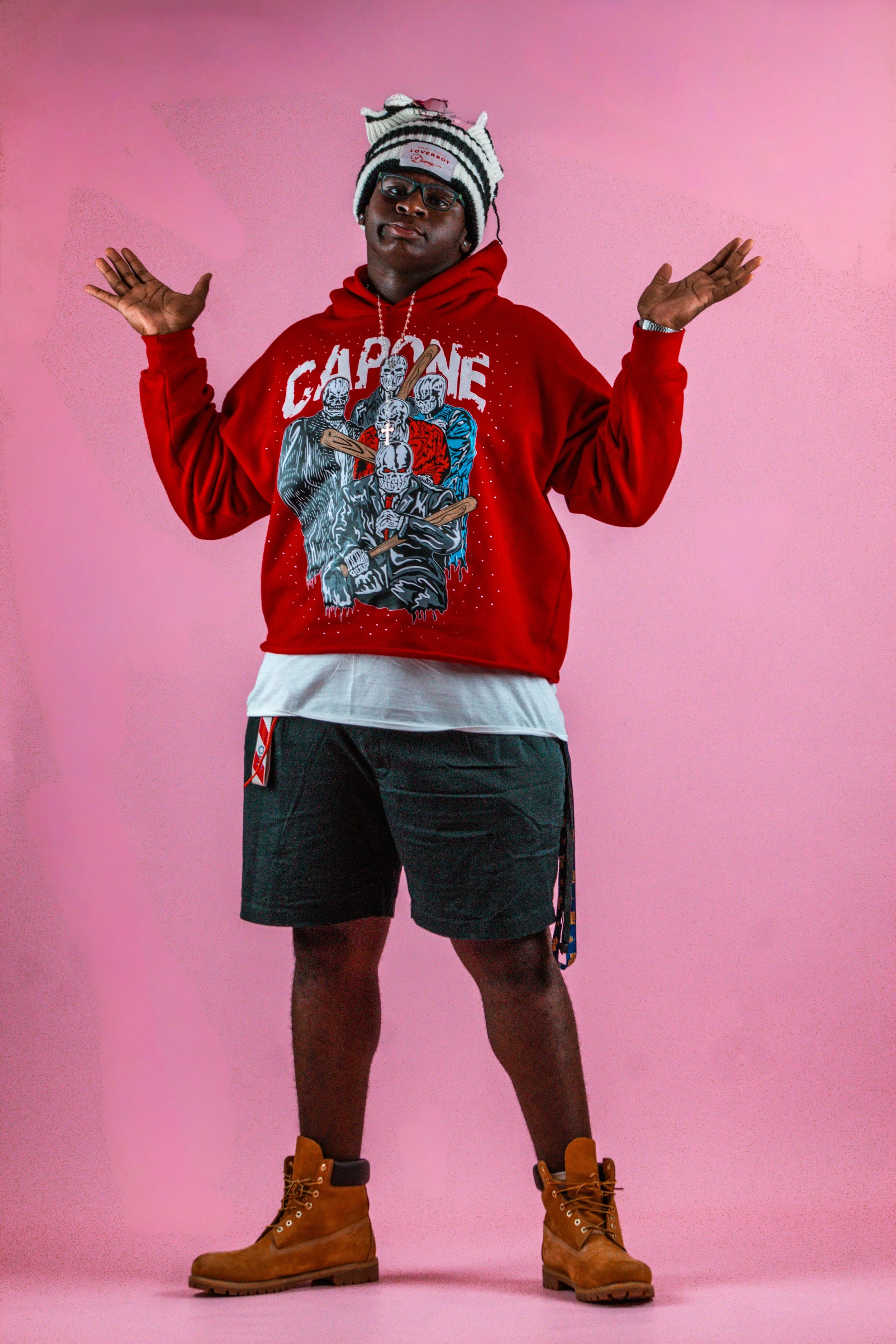 Person standing with hands raised against a pink background, wearing a red hoodie, black shorts, tan boots, glasses, and a striped hat.