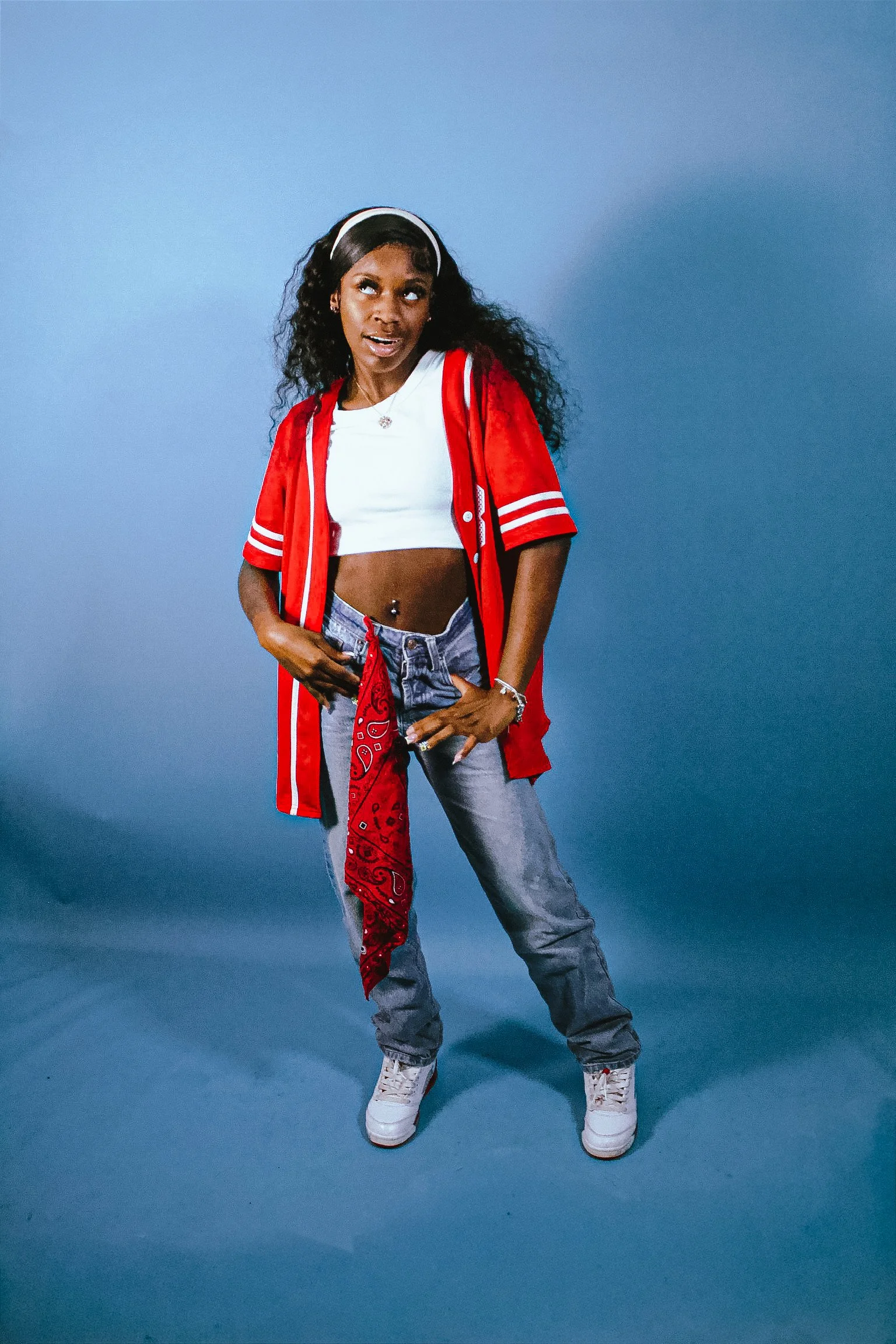 A woman using a pose commonly associated with rap or hip-hop culture, standing against a blue background, wearing a red and white varsity jacket, white crop top, gray jeans, and white sneakers, with long curly hair and a white headband.