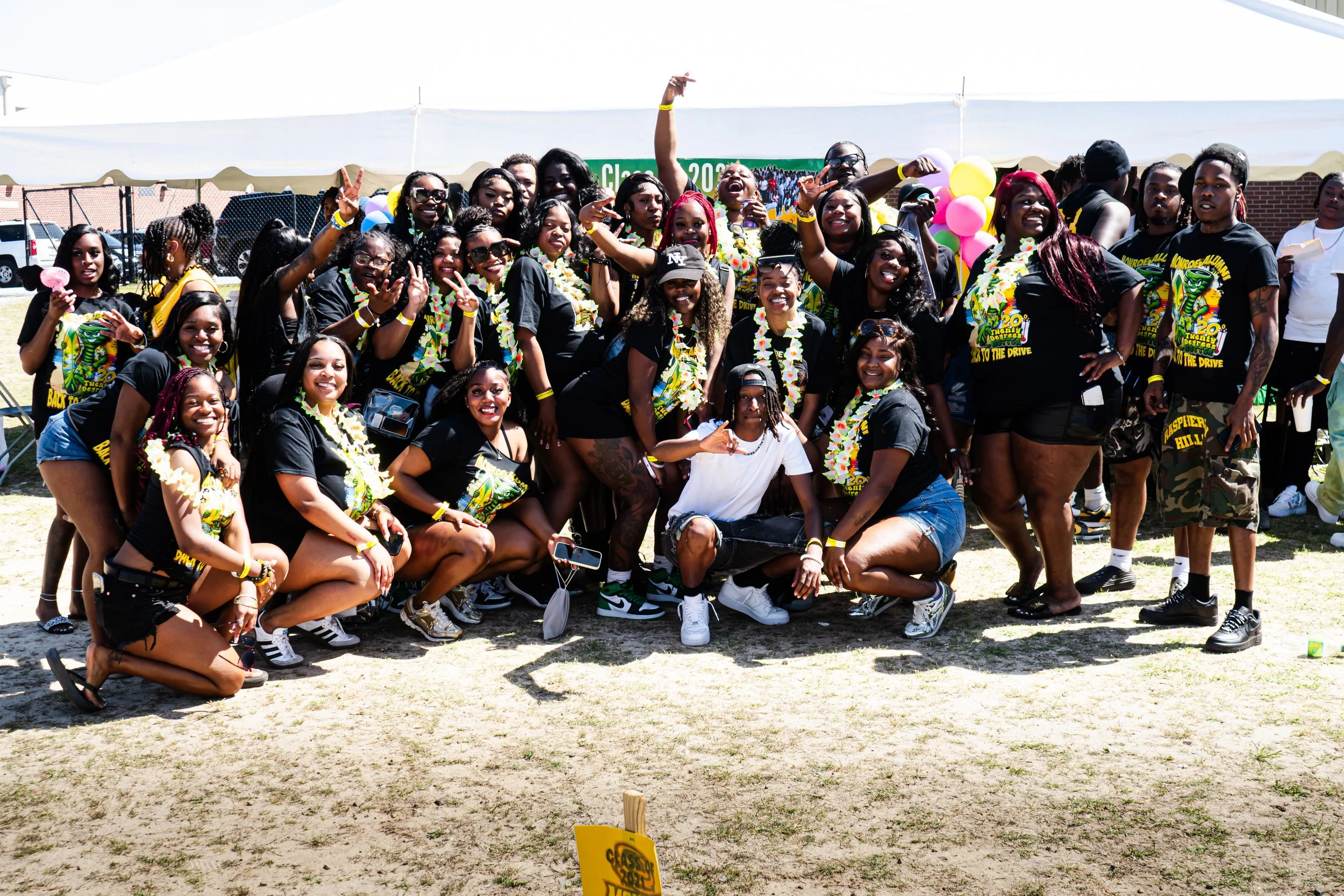 A group of people, mostly women, smiling and posing together at an outdoor event under a white canopy tent. Many are wearing black T-shirts with colorful graphics, leis, and yellow wristbands. Some are making peace signs and other hand gestures. Ther