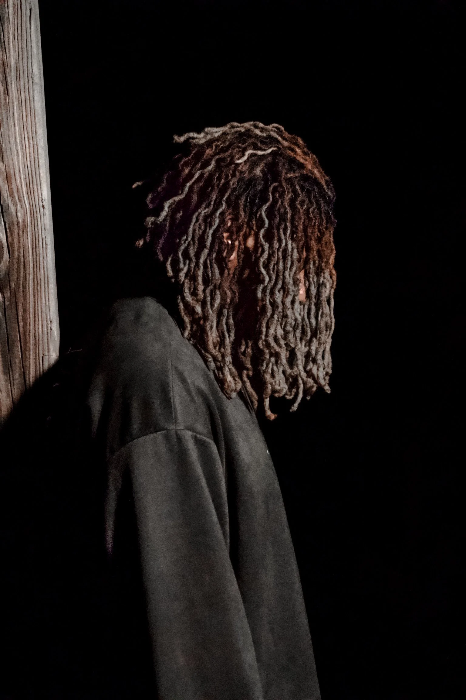 Person with long dreadlocks wearing a black jacket standing near a wooden post at night.