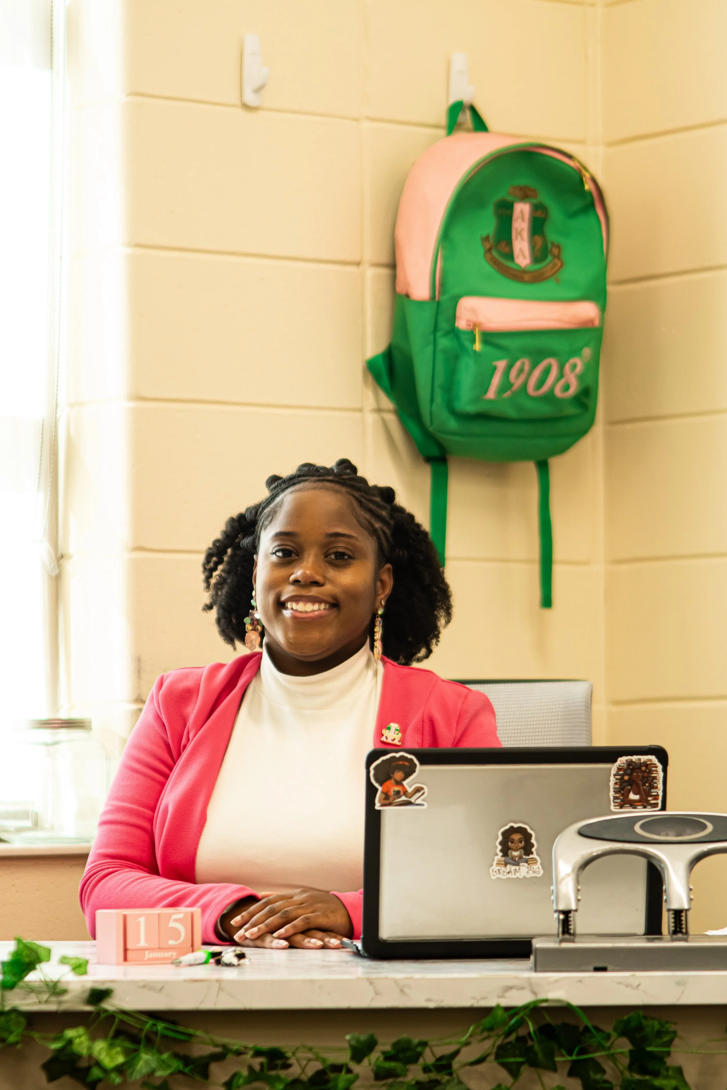 A woman with dark curly hair and earrings smiling while sitting at a desk with a laptop. There is a green and pink backpack hanging on the wall behind her with the numbers '1908' on it. The desk has a pink calendar, some decorations, and stickers on 