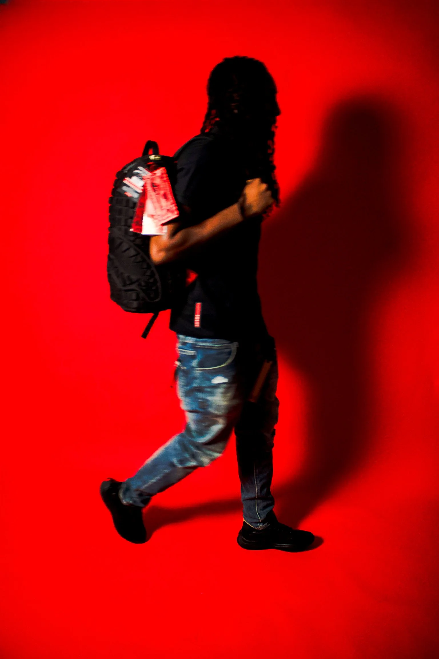 Person walking with a backpack against a red background, dressed in casual clothing.