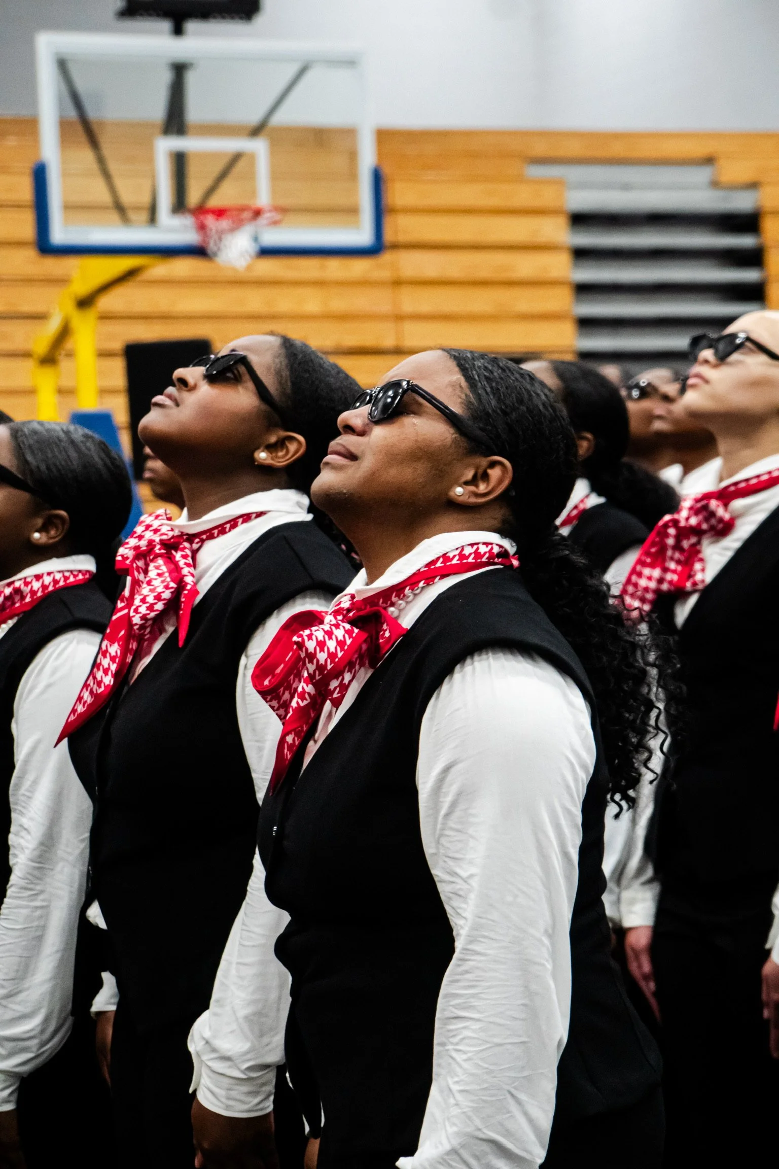 A group of women wearing white shirts, black vests, red and white patterned scarves, and sunglasses standing inside a gymnasium with a basketball hoop and wooden walls.