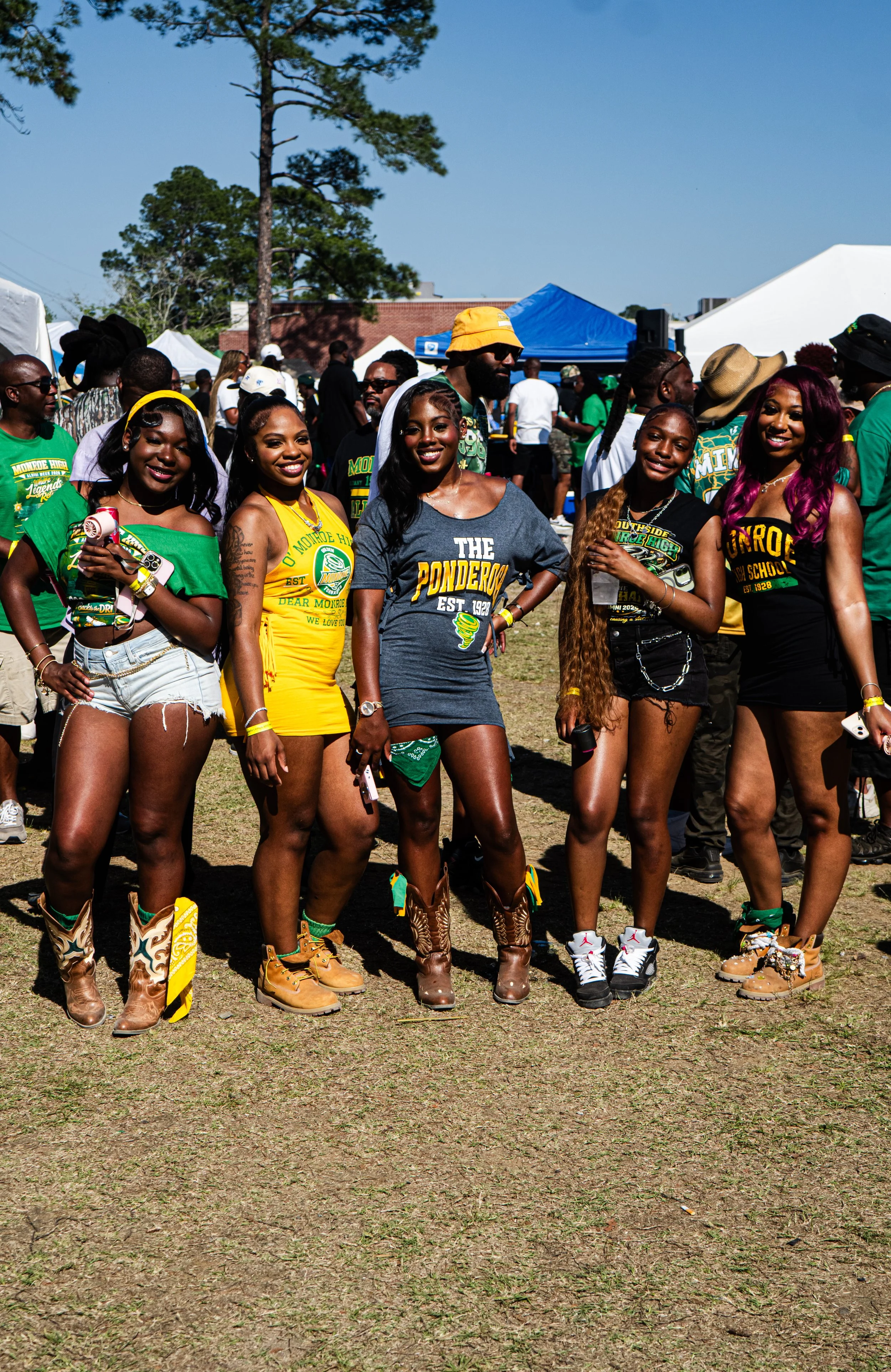 Group of people gathered outdoors at a festival or event, wearing colorful and casual clothing, some holding drinks, with tents and trees in the background.