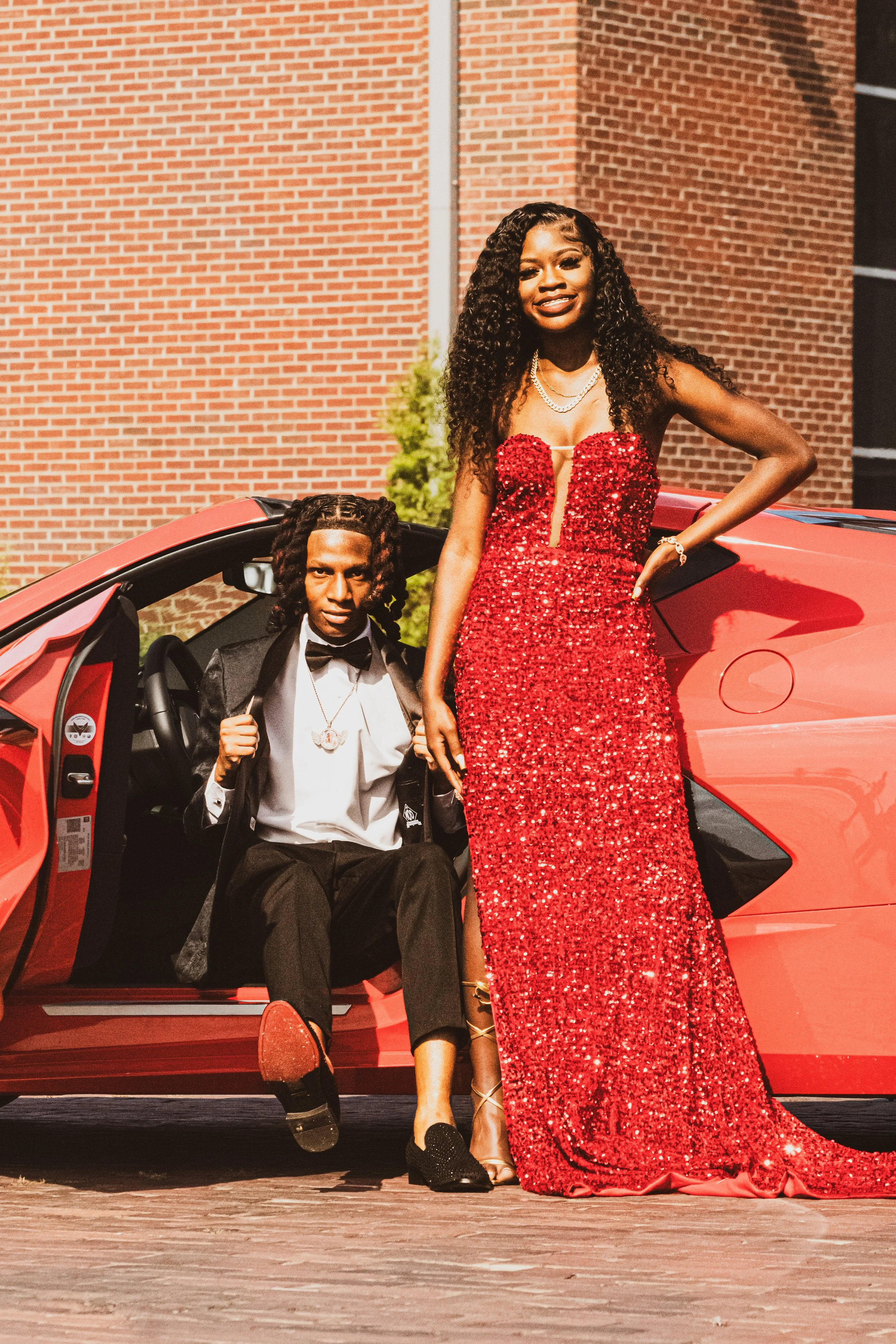 A young woman in a red sequined gown standing next to a red sports car, with a young man in a suit sitting in the car, both posing outdoors in front of a brick building.