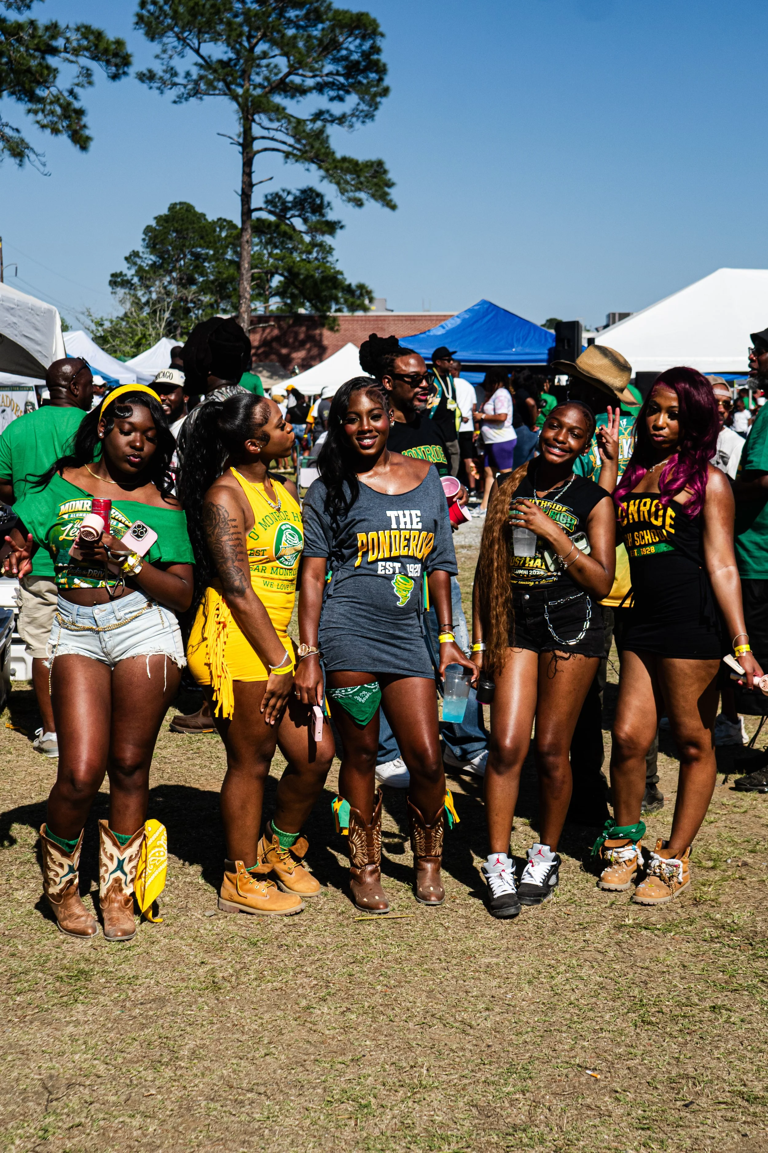 A group of five women standing outdoors at a festival or fair, wearing casual and colorful outfits with boots, surrounded by tents and other people in the background.
