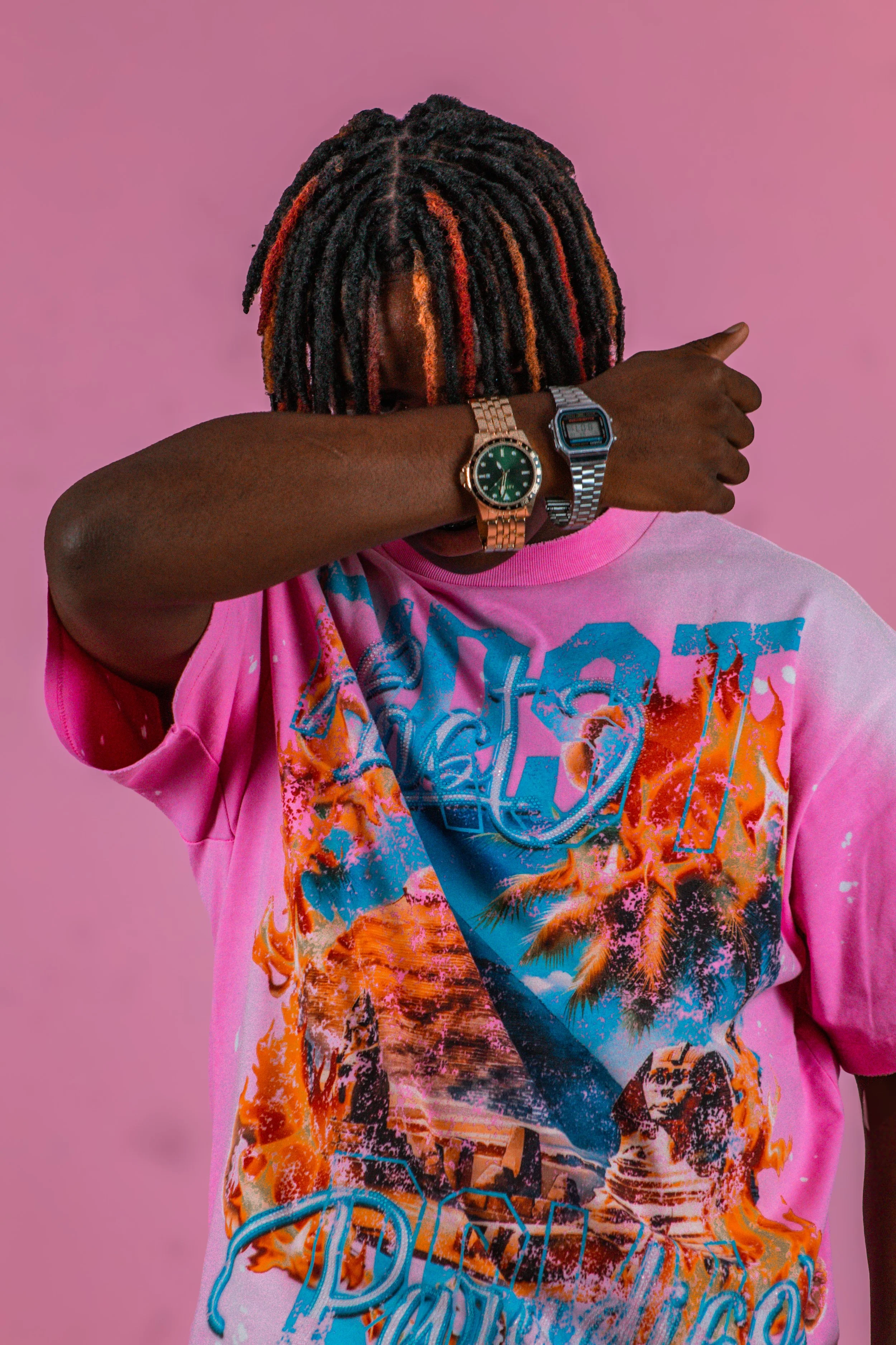 Person with multicolored dreadlocks, wearing a pink graphic T-shirt, a gold watch, and a silver digital watch, standing against a pink background.