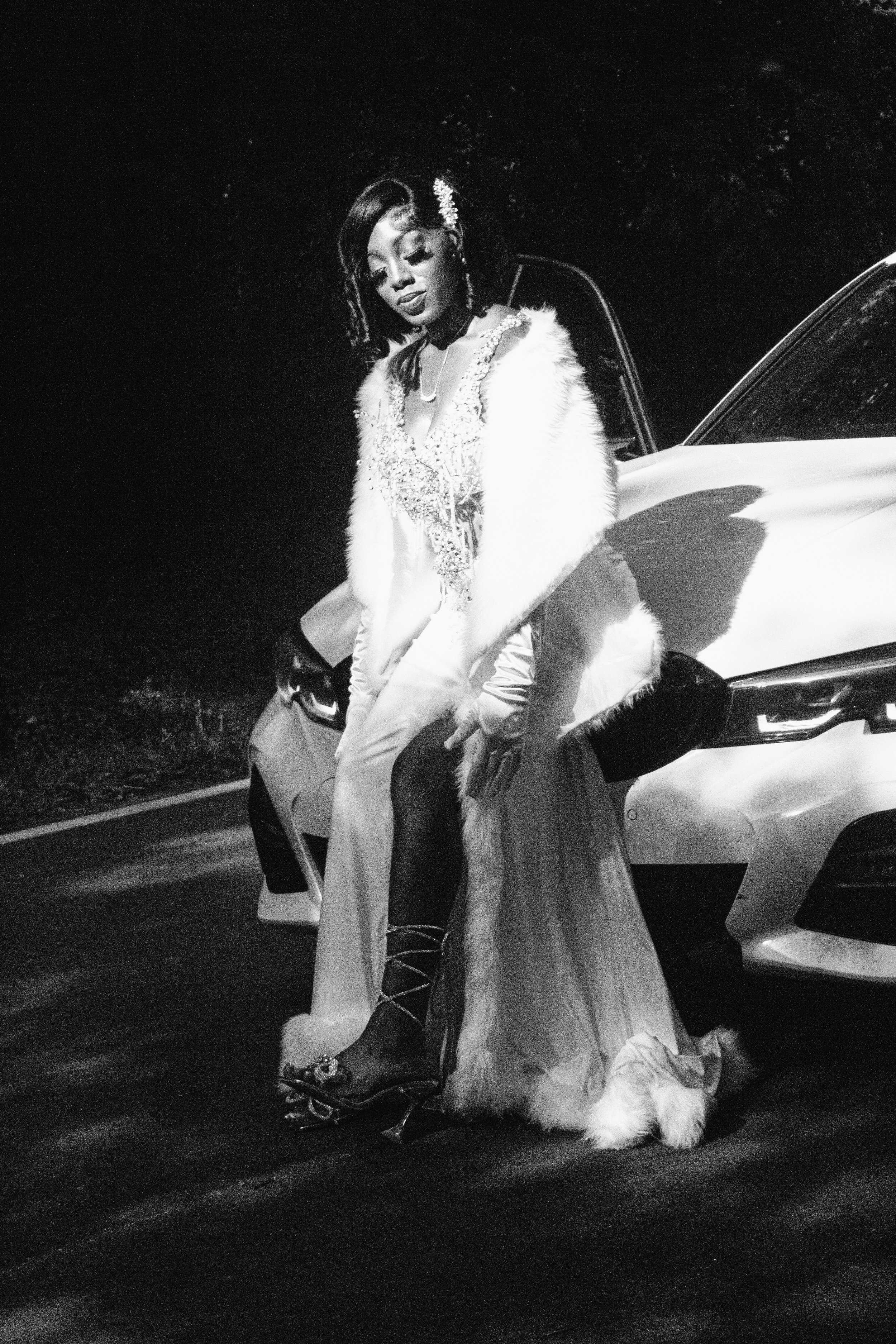 Black and white image of a woman in an elegant dress with a fur stole, standing next to a luxury car at night.