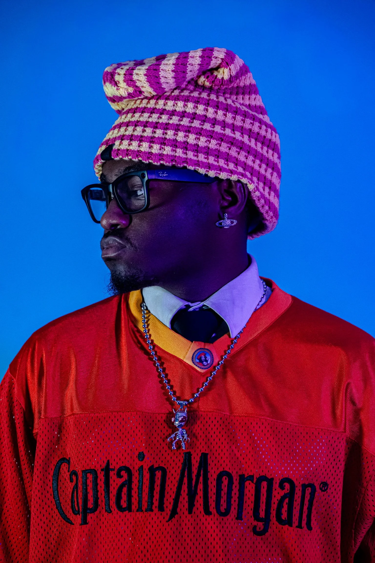 A person wearing a pink and yellow knitted beanie, black glasses, a red sports jersey with 'Captain Morgan' written on it, and a layered necklace with a pendant, set against a blue background.