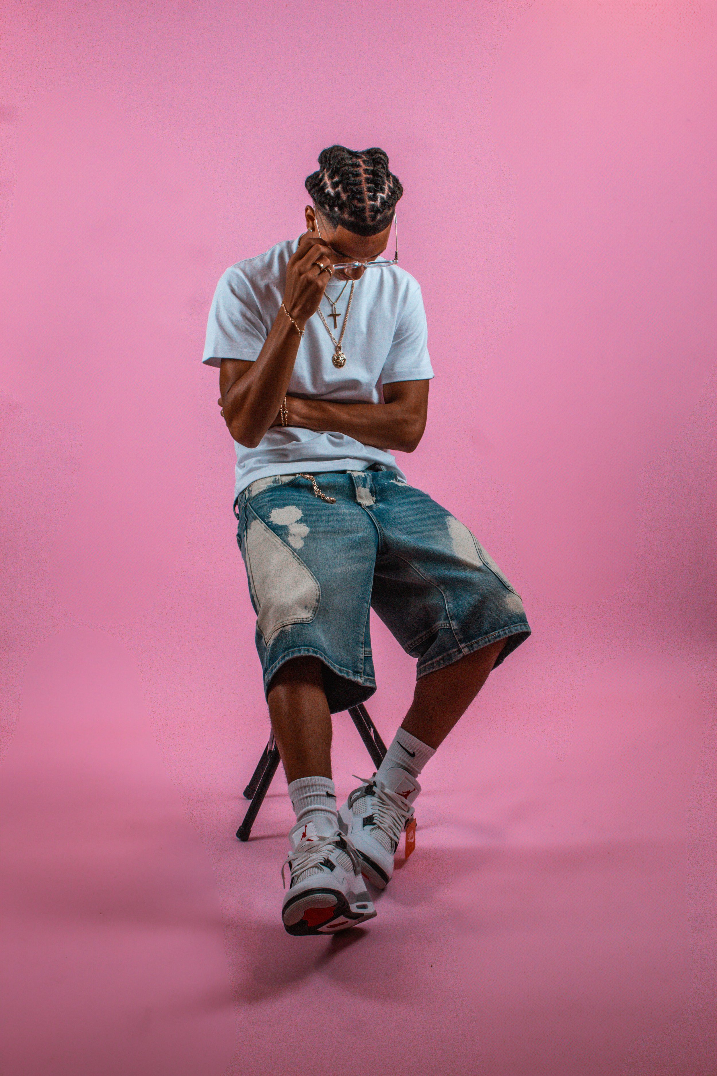 Young man with braids, wearing a gray t-shirt, baggy denim shorts, white sneakers, white socks, and jewelry, sitting on a black stool against a pink background, looking down while adjusting glasses