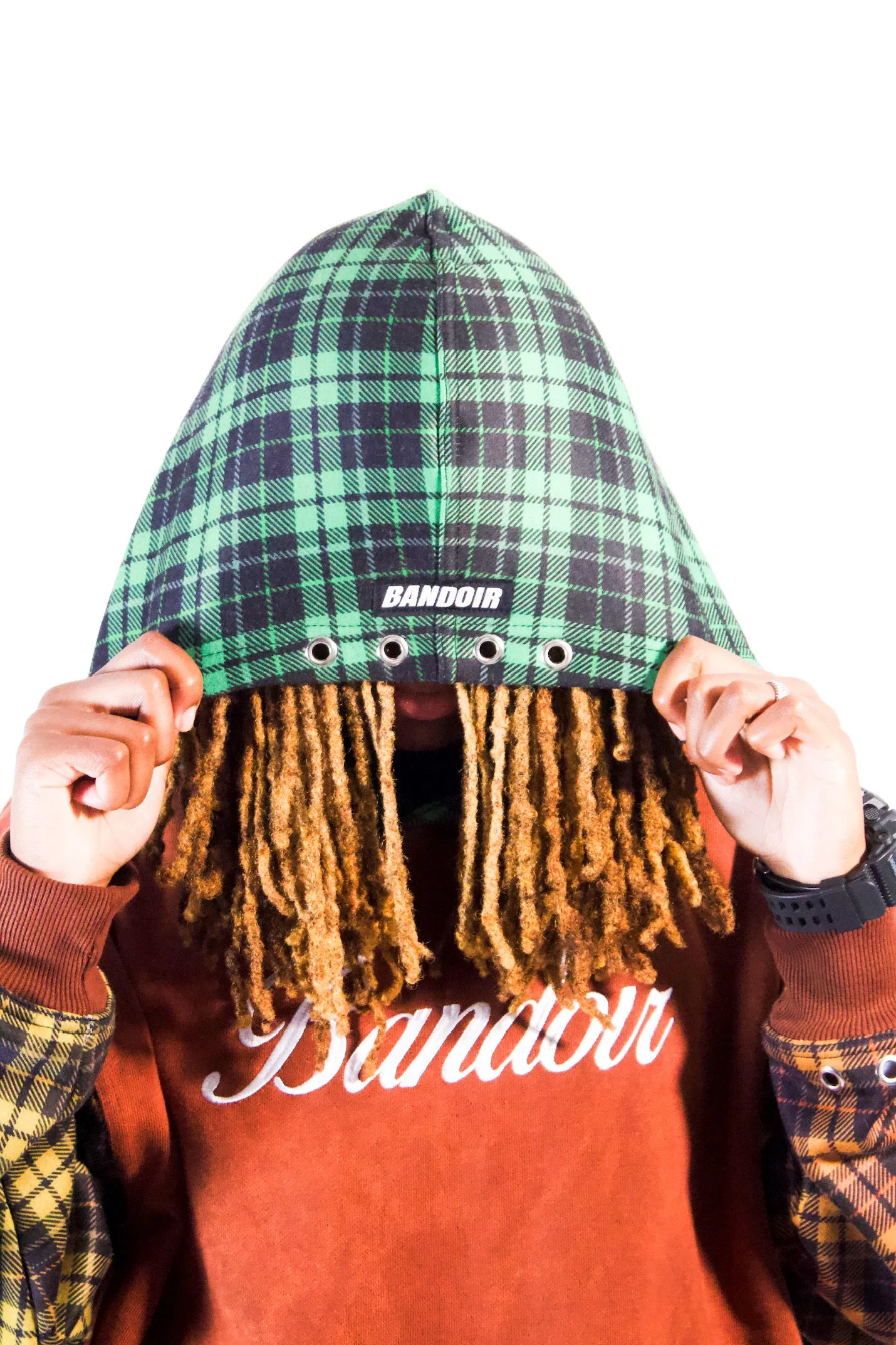 Person holding a green tartan-patterned hat labeled 'BANDOIR' in front of their face, wearing a maroon hoodie with white script, plaid shirt cuffs, and a wristwatch.
