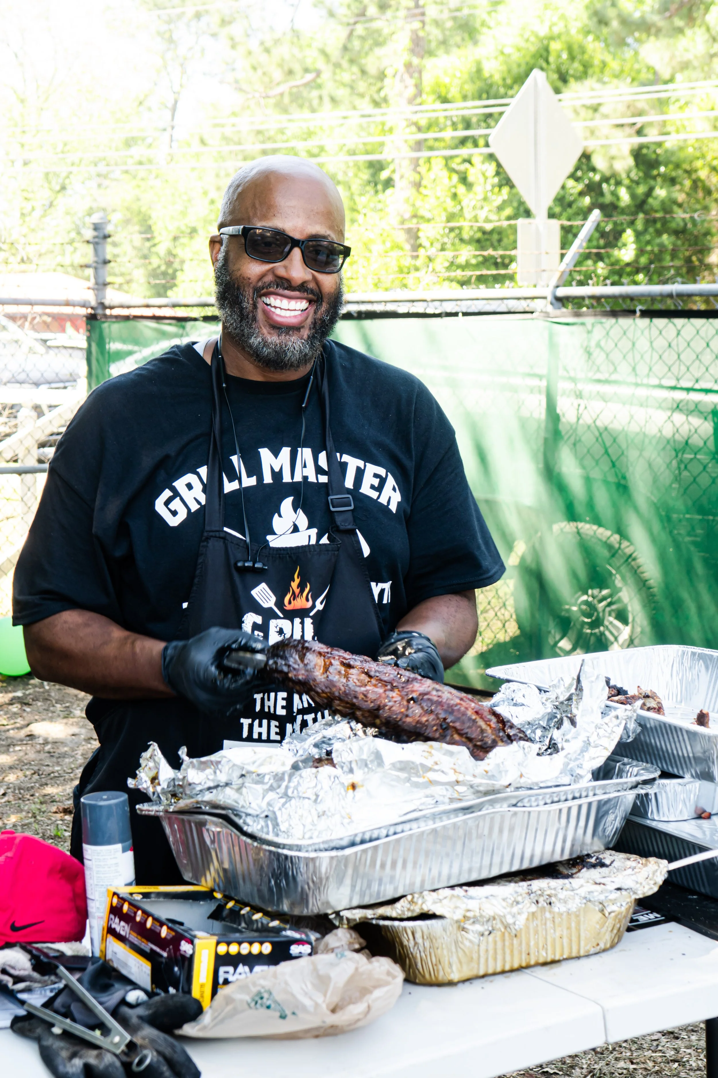 A man with dark sunglasses, a beard, and a black t-shirt with white text, smiling and holding a large cooked meat, standing at an outdoor barbecue table with aluminum trays of food, surrounded by trees and a green fence.