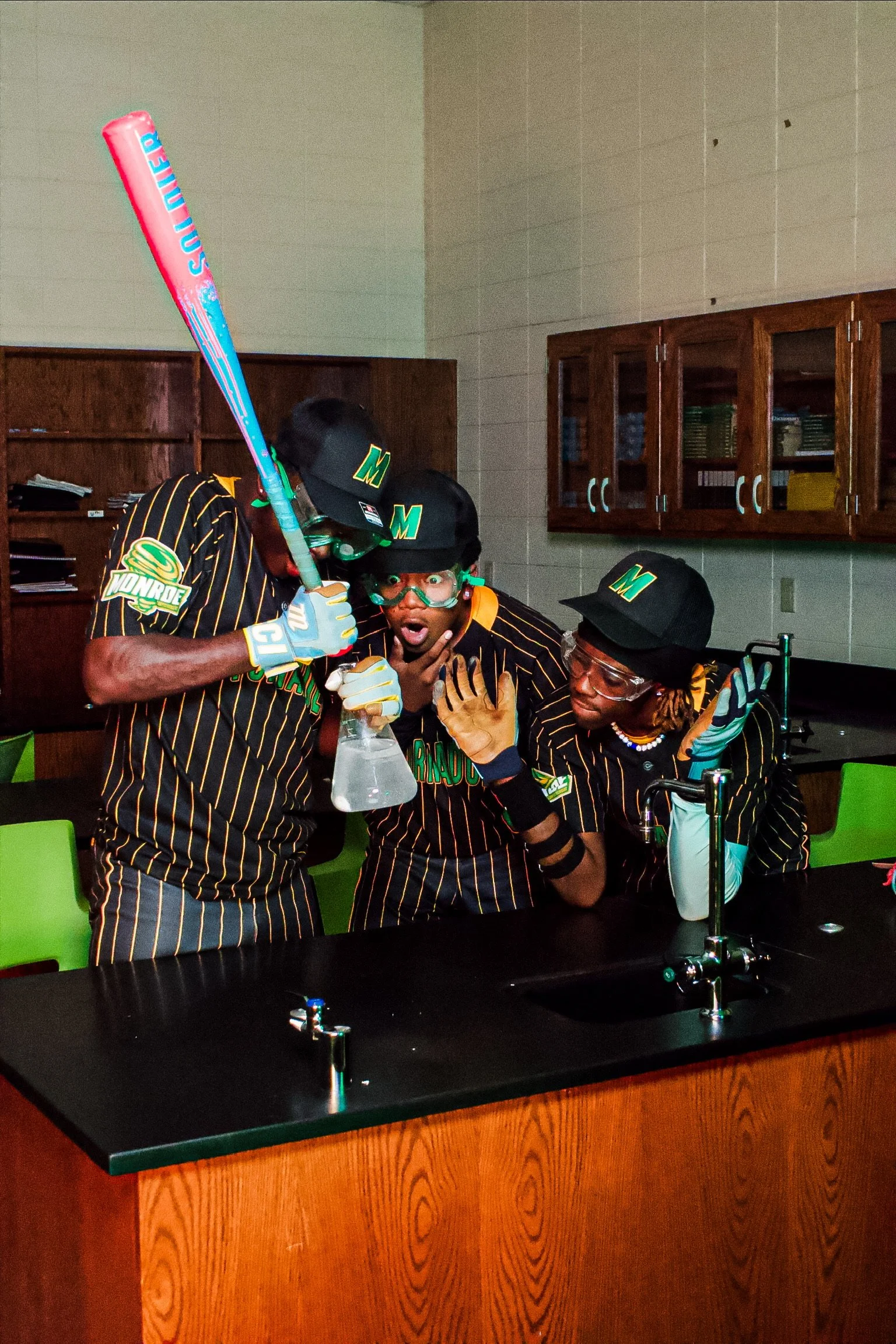 Group of four young men dressed in baseball uniforms, wearing caps with a green 'M' logo, gathered around a school science lab table with a glass bottle and flask, in a classroom setting.