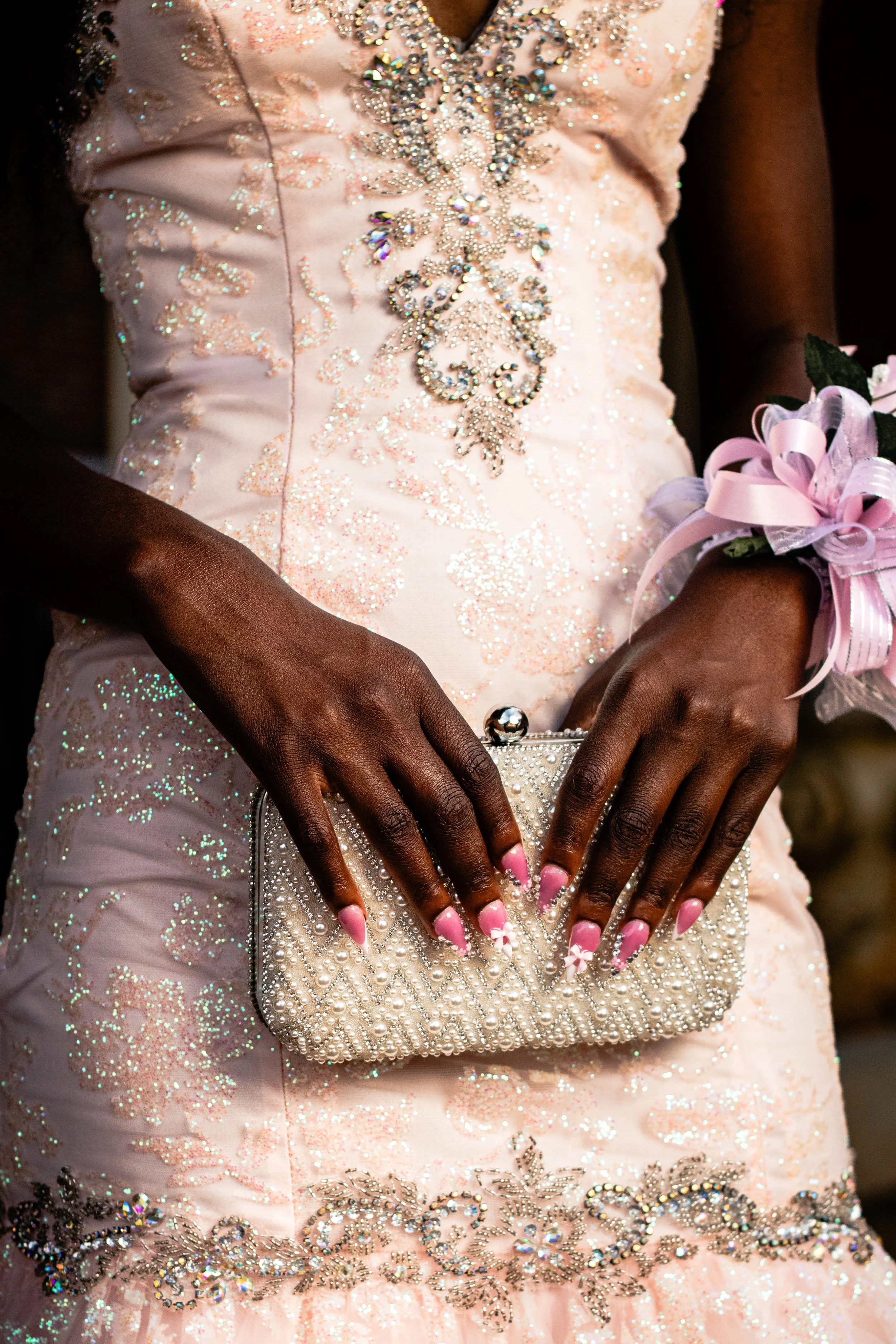 Close-up of a pink dress decorated with silver and iridescent beads and sequins. A person with dark skin is holding a pearl-embellished clutch bag with a bejeweled clasp. They are wearing a wrist corsage with pink ribbons and a flower. Their nails ar