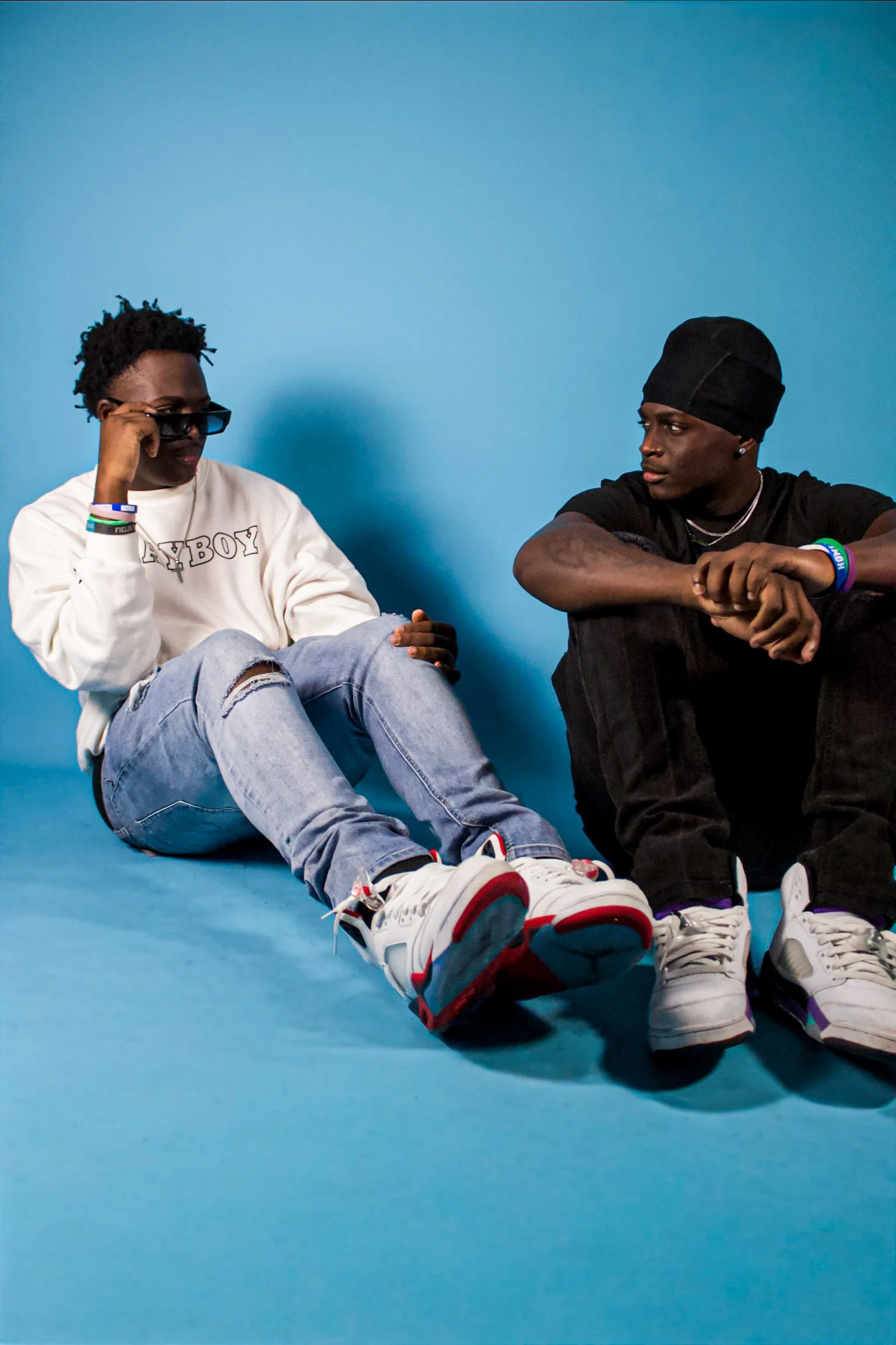Two young men sitting on a blue floor against a blue wall, one wearing a white sweatshirt and ripped jeans, the other wearing a black beanie and black t-shirt, both wearing white sneakers and colorful wristbands, engaged in casual conversation.