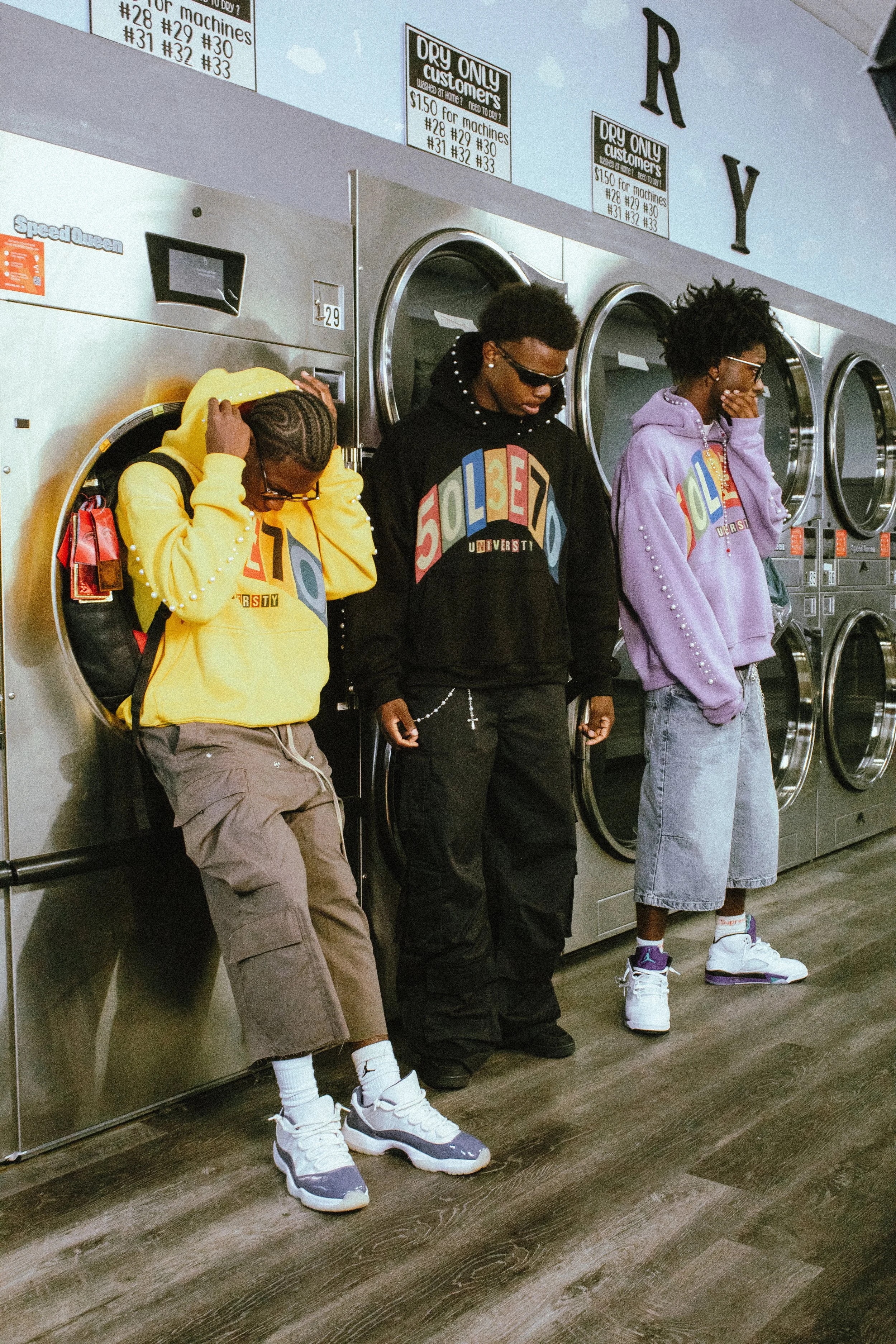 Three young men standing in a laundromat, wearing colorful hoodies and sneakers, with washing machines in the background.