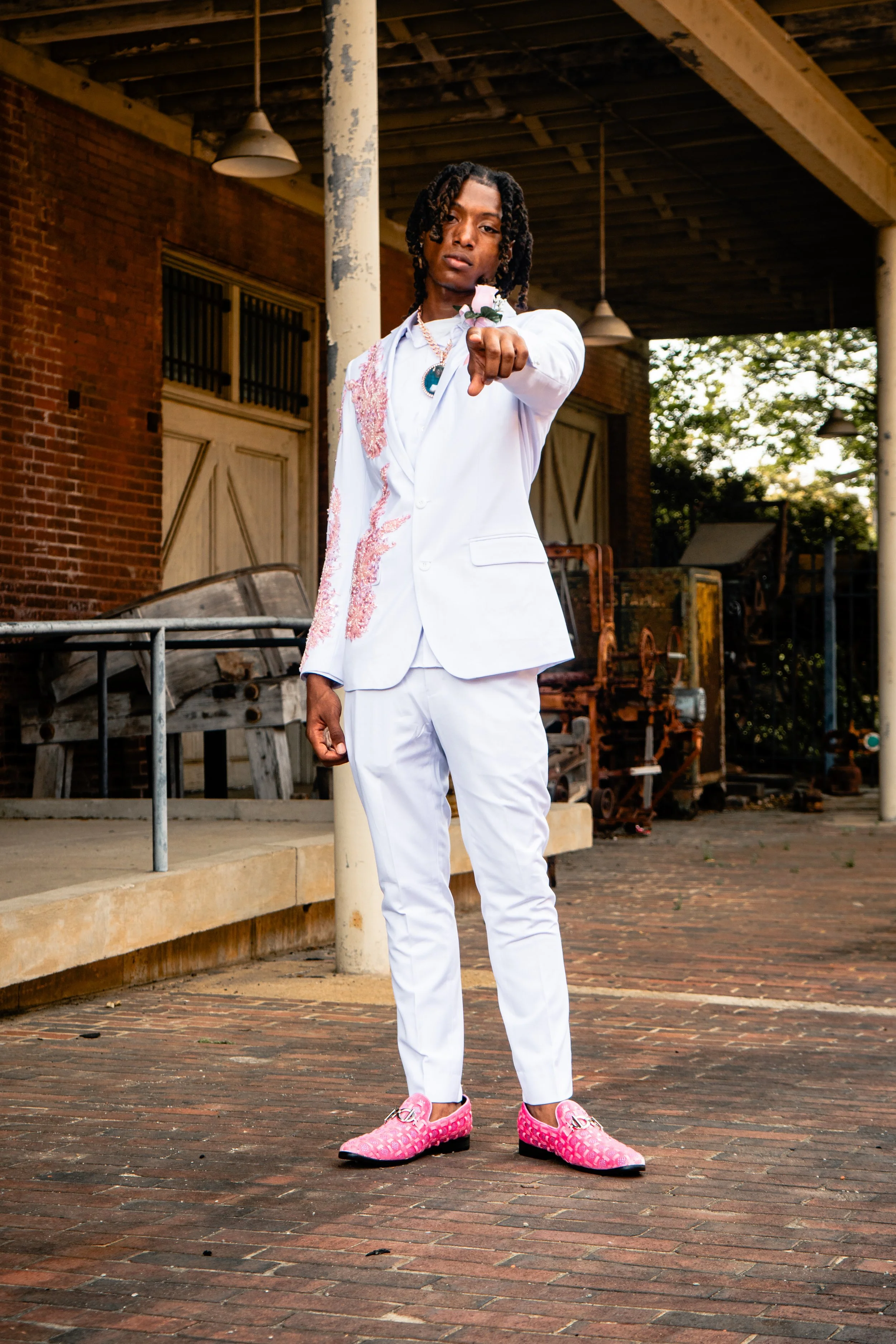 Young man with dreadlocks wearing white suit, pink shoes, and a flower on his wrist, standing outdoors on a brick sidewalk, pointing towards the camera.