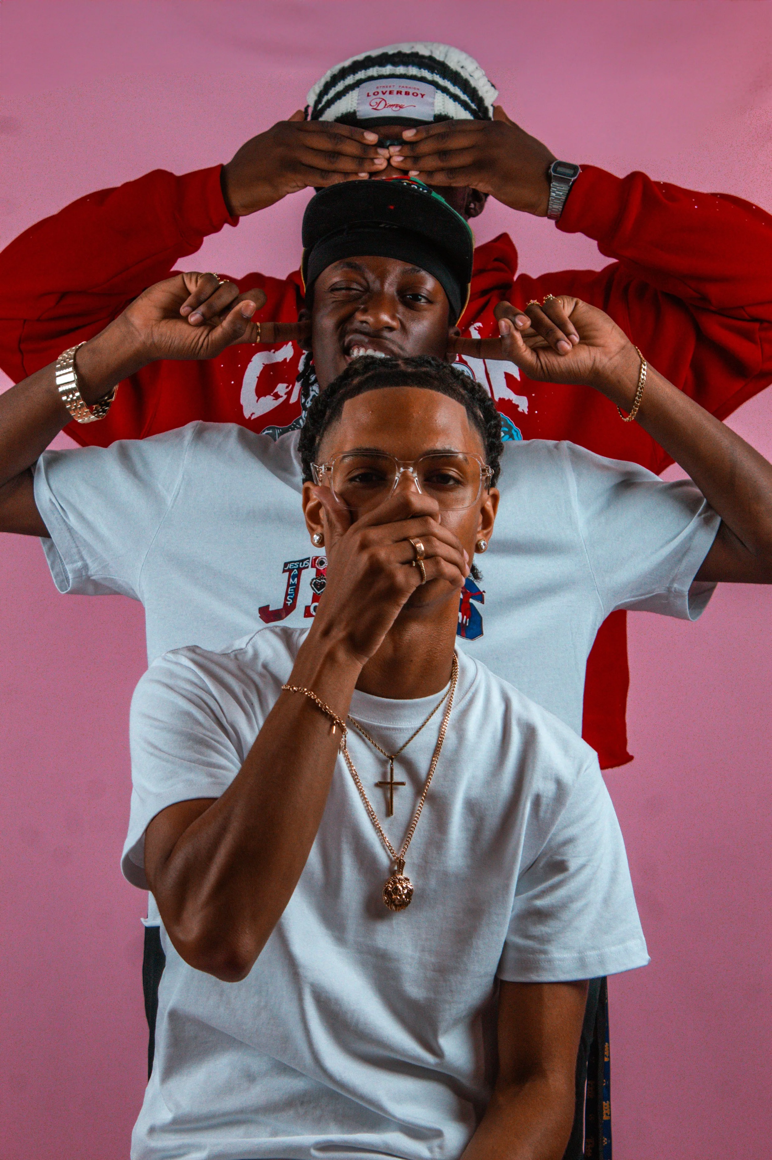 Two young men posing playfully against a pink background. One in a white T-shirt and glasses, covering his mouth with his hand, the other in a red hoodie with a black cap, pulling on his ears and squinting.