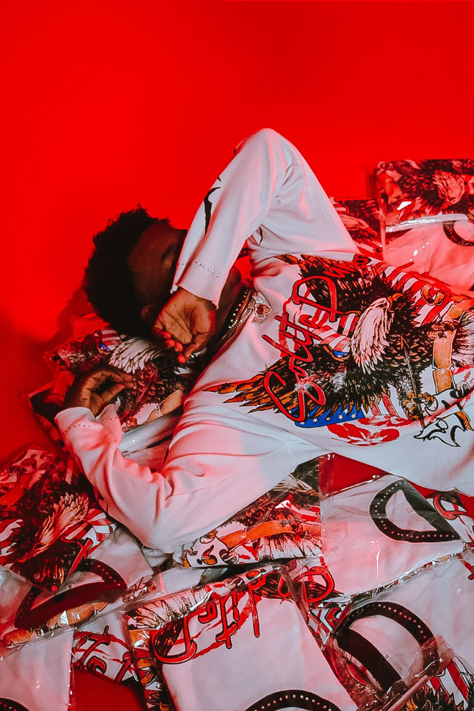 A person laying down on a surface covered with a colorful, patterned fabric, posing with their eyes closed, wearing a white jacket with a graphic design and text, against a vivid red background.