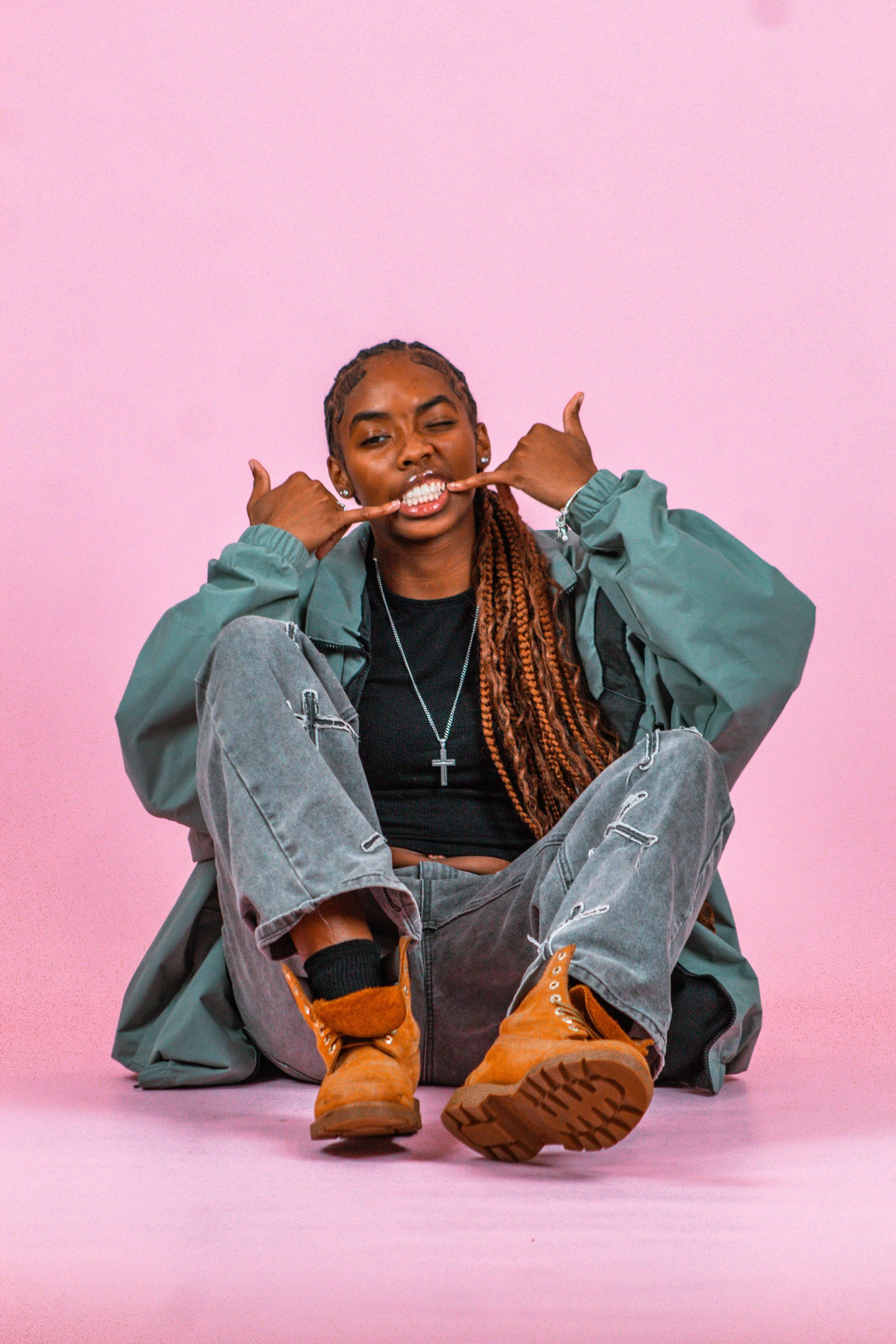 A young woman with braided hair, wearing a black shirt, gray jeans, an oversized gray jacket, orange boots, and a cross necklace, sitting against a pink background, winking and making a playful gesture with her hands.