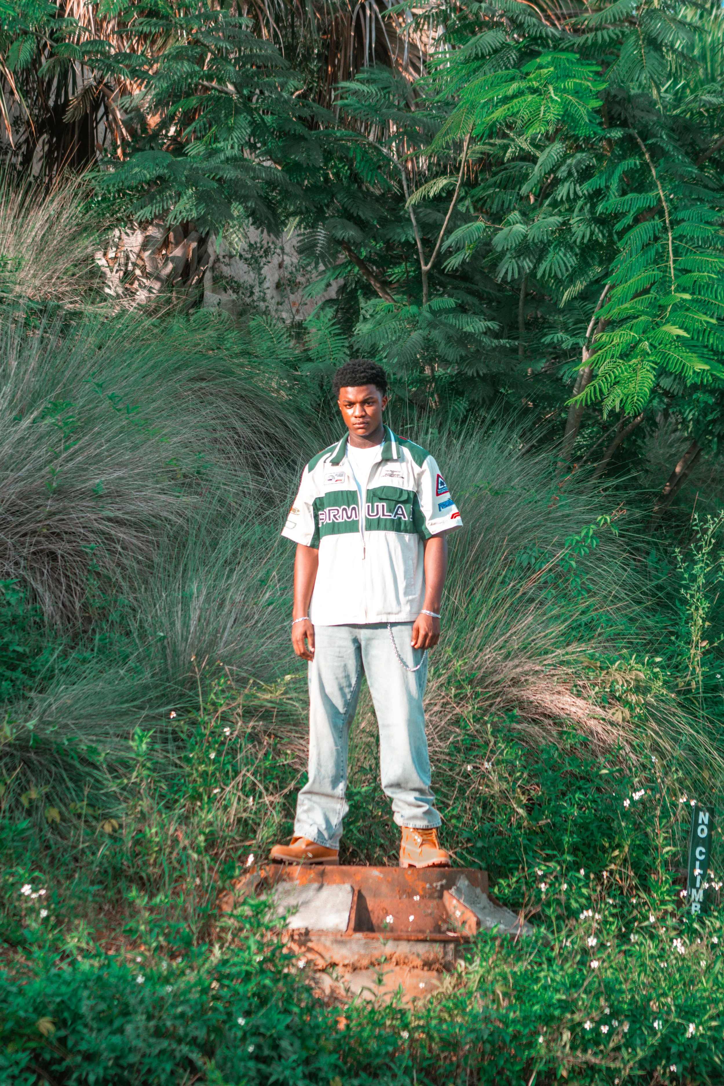 A young man standing outdoors among green plants and trees. He is wearing a white and green racing jacket, gray baggy pants, and orange shoes, with a serious expression on his face.