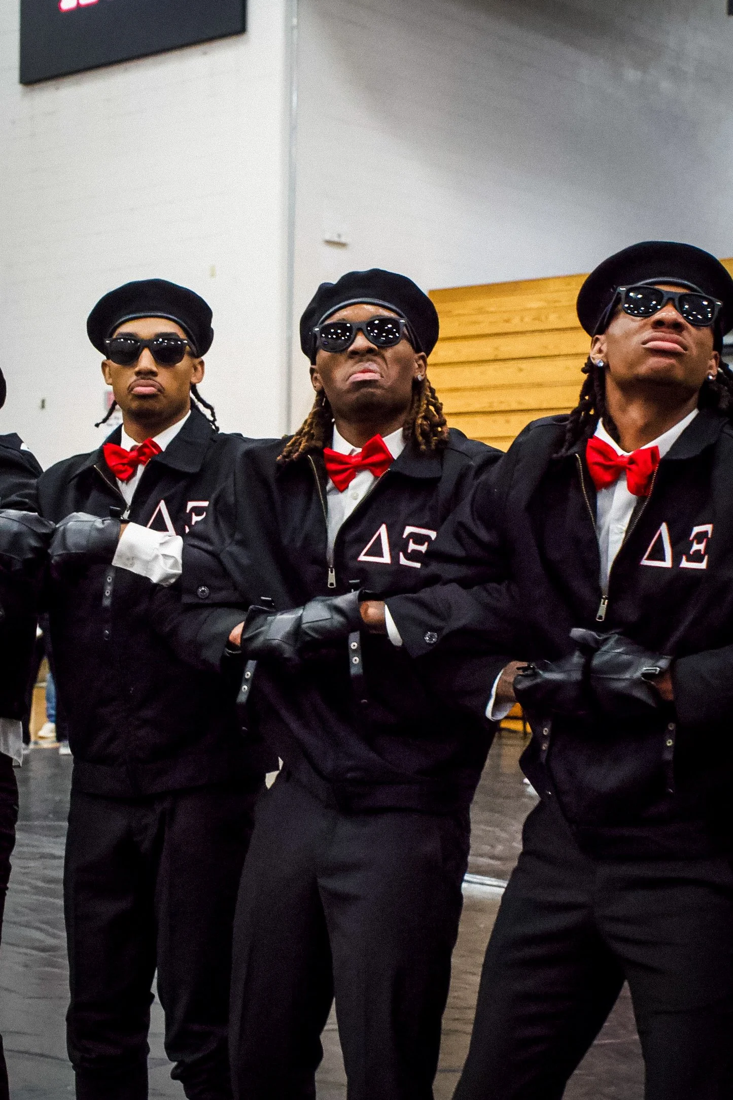 Three individuals dressed in black suits and red bow ties, wearing black sunglasses and black berets, standing with arms crossed. The background features a wood-paneled wall and gymnasium setting.