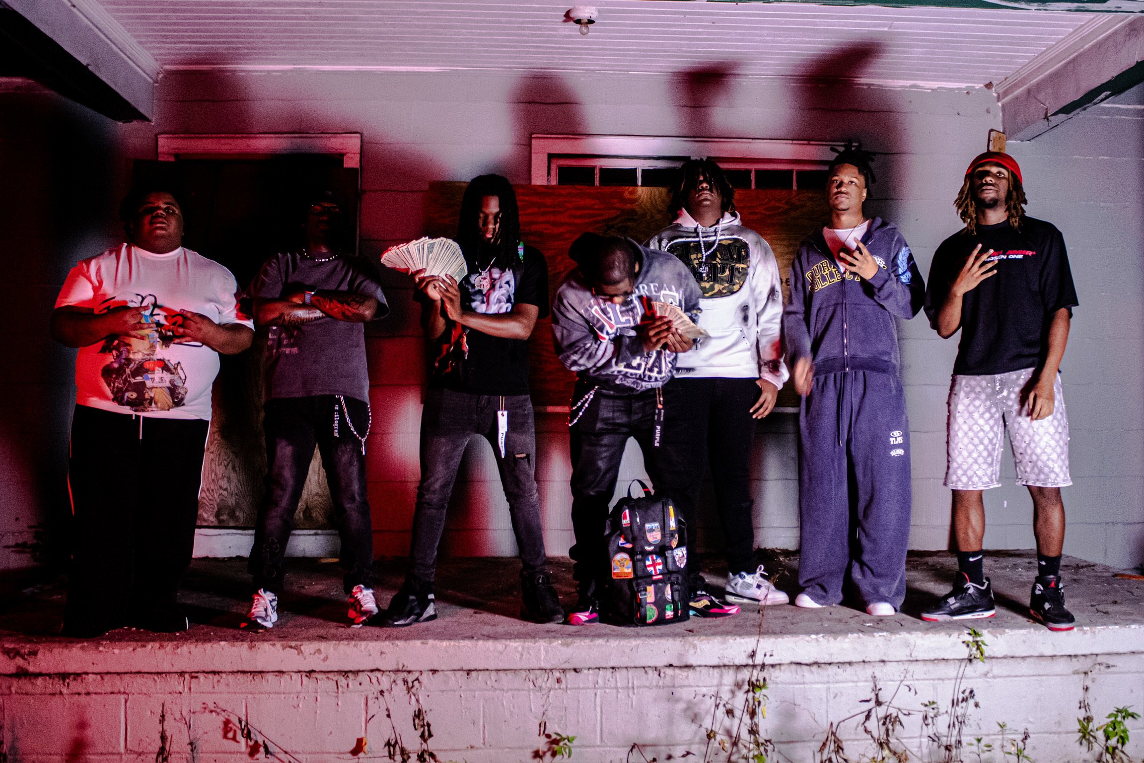 Group of seven young men standing outdoors at night, some holding money and electronic devices, one with a backpack, in front of a wooden wall. They are dressed casually in hoodies, t-shirts, and shorts.