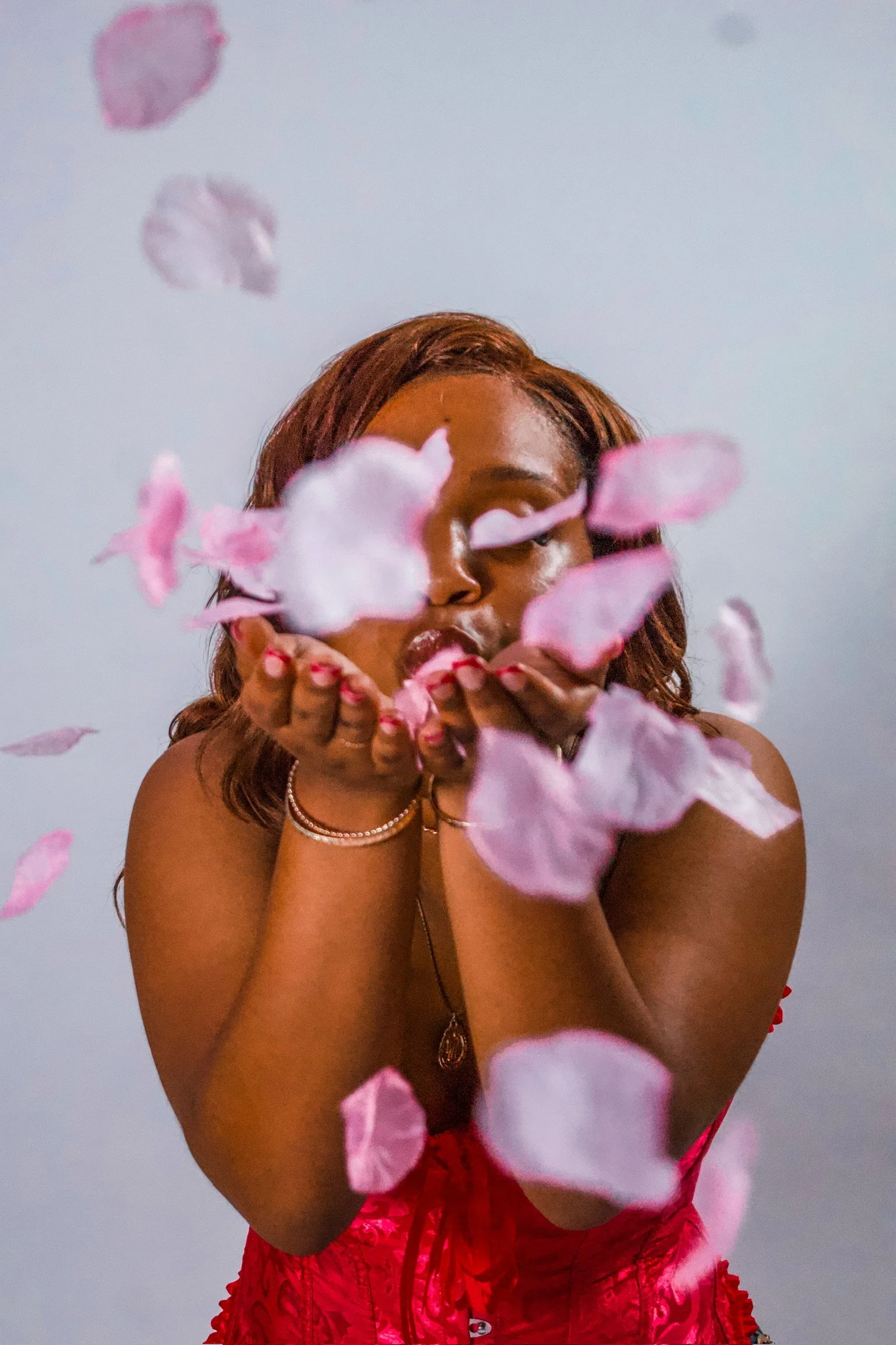 A person with long, wavy brown hair and tan skin blowing pink flower petals towards the camera. They are wearing a red outfit with ruffles and jewelry, and are standing in front of a plain wall.