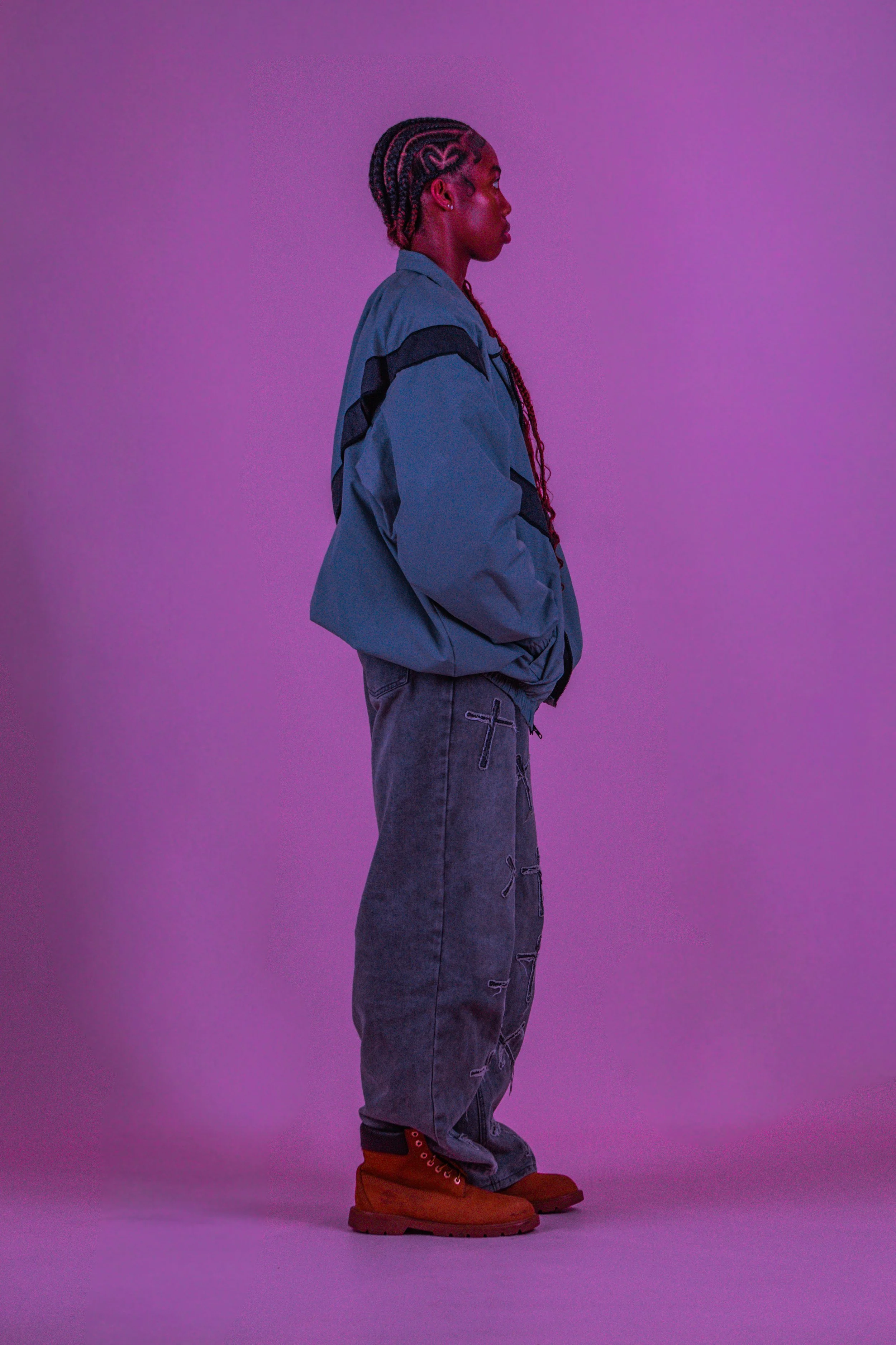 Side profile of a person with braided hair, wearing a blue jacket, gray ripped jeans, and brown boots against a purple background.