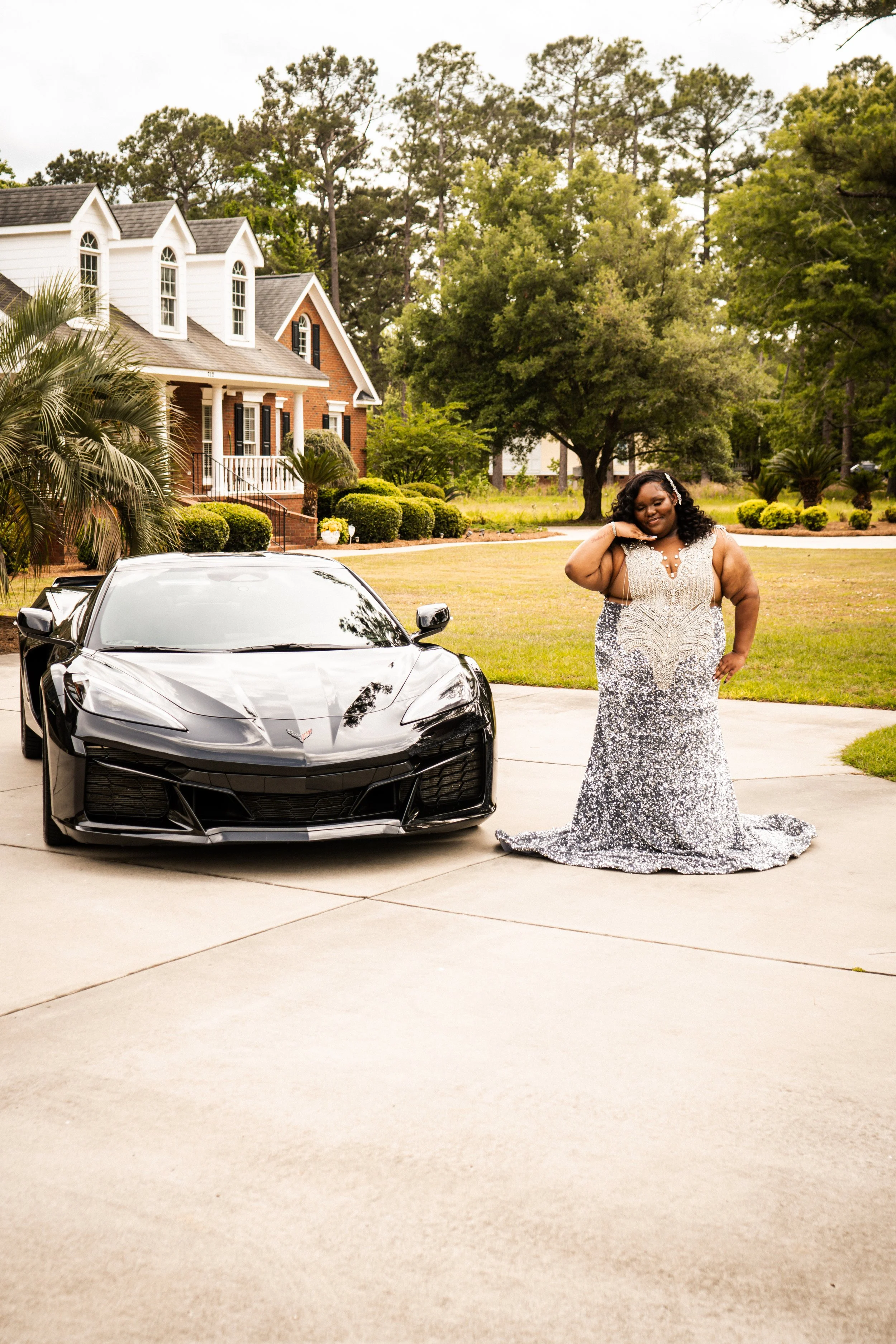 A woman in a silver, sequined, evening gown is standing outside a large house with a porch, surrounded by trees, next to a black sports car, with her hand resting on her cheek, smiling.