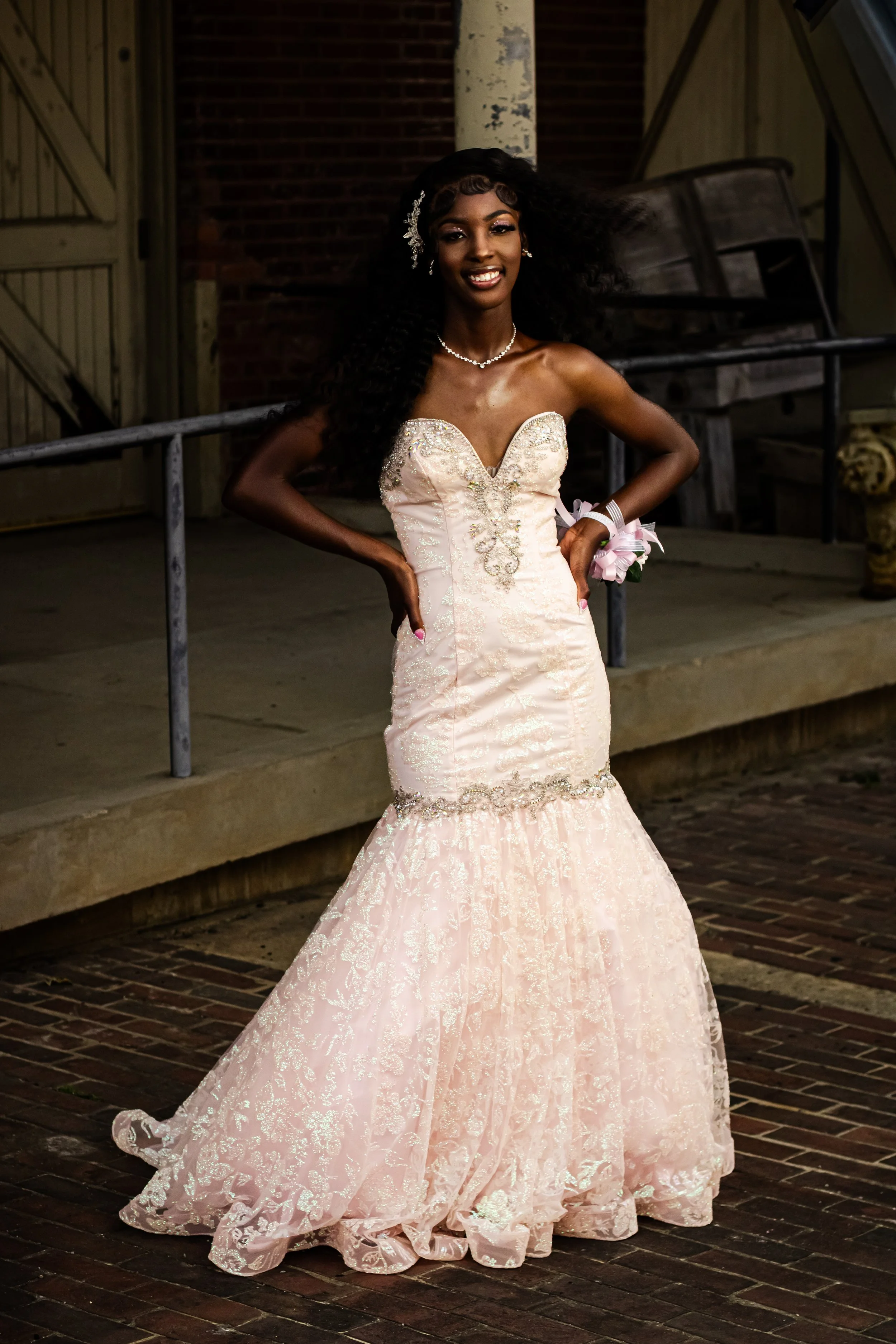 A woman in a strapless, light pink wedding gown with lace detailing, standing outdoors on a brick pathway with her hands on her hips, smiling at the camera.