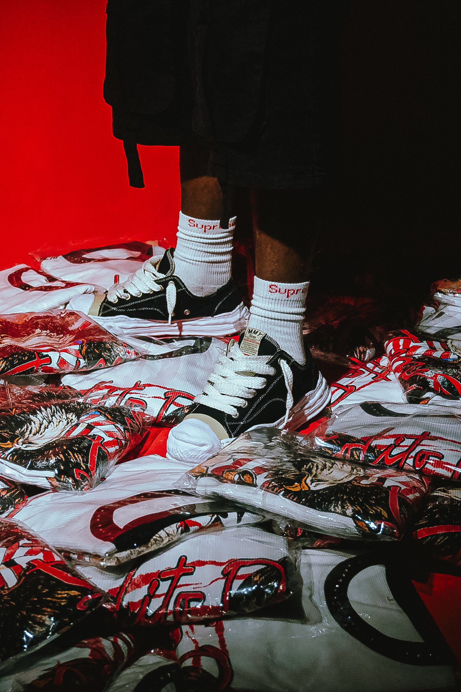 A person standing on a floor covered with red, white, and black plastic bags with a graphic design, wearing black shorts, white socks with red text, and black sneakers, with a red wall in the background.