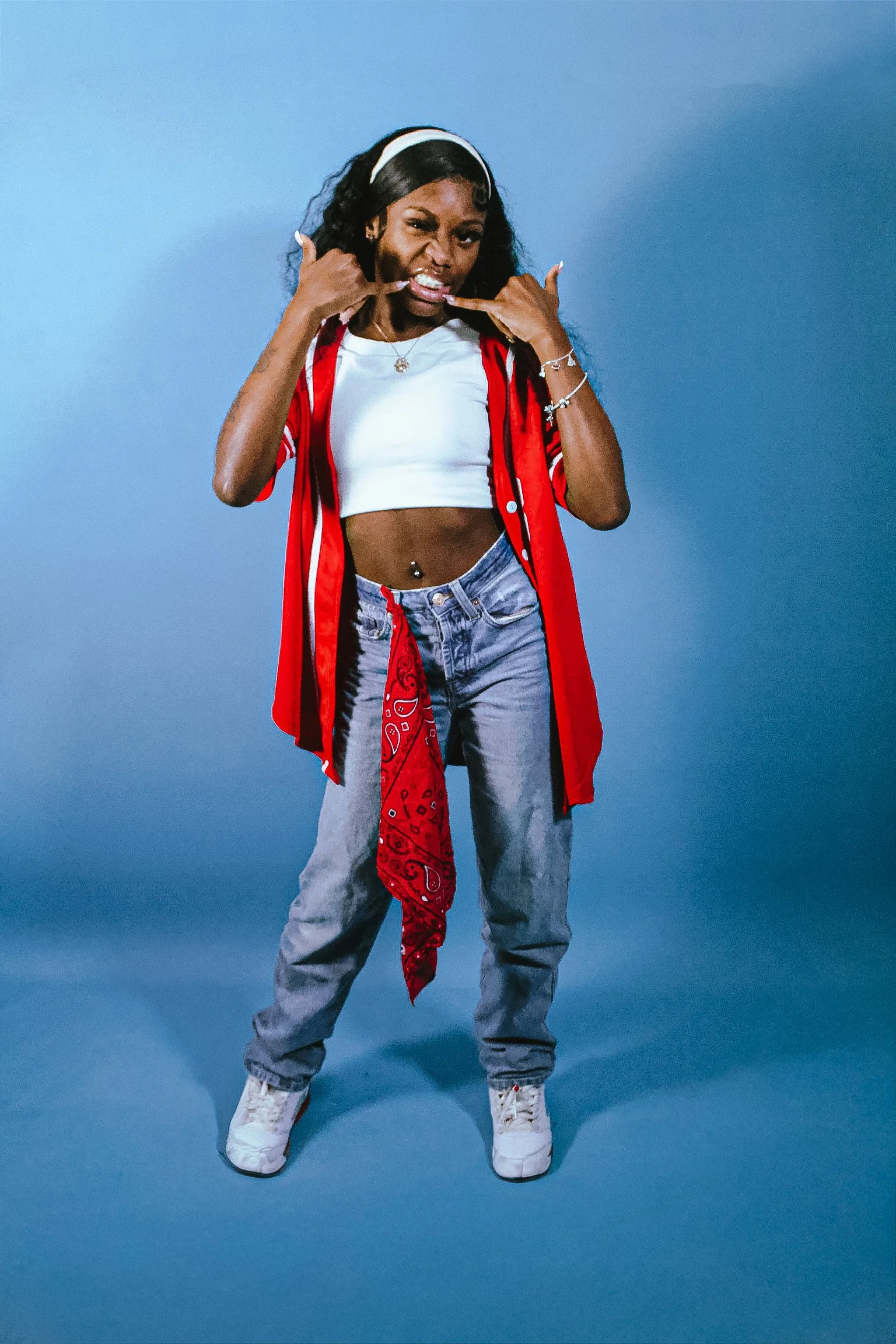 A woman standing on a blue background, wearing a white crop top, a red open shirt, light blue jeans, white sneakers, and accessorized with jewelry, making a playful gesture with her hands near her face.
