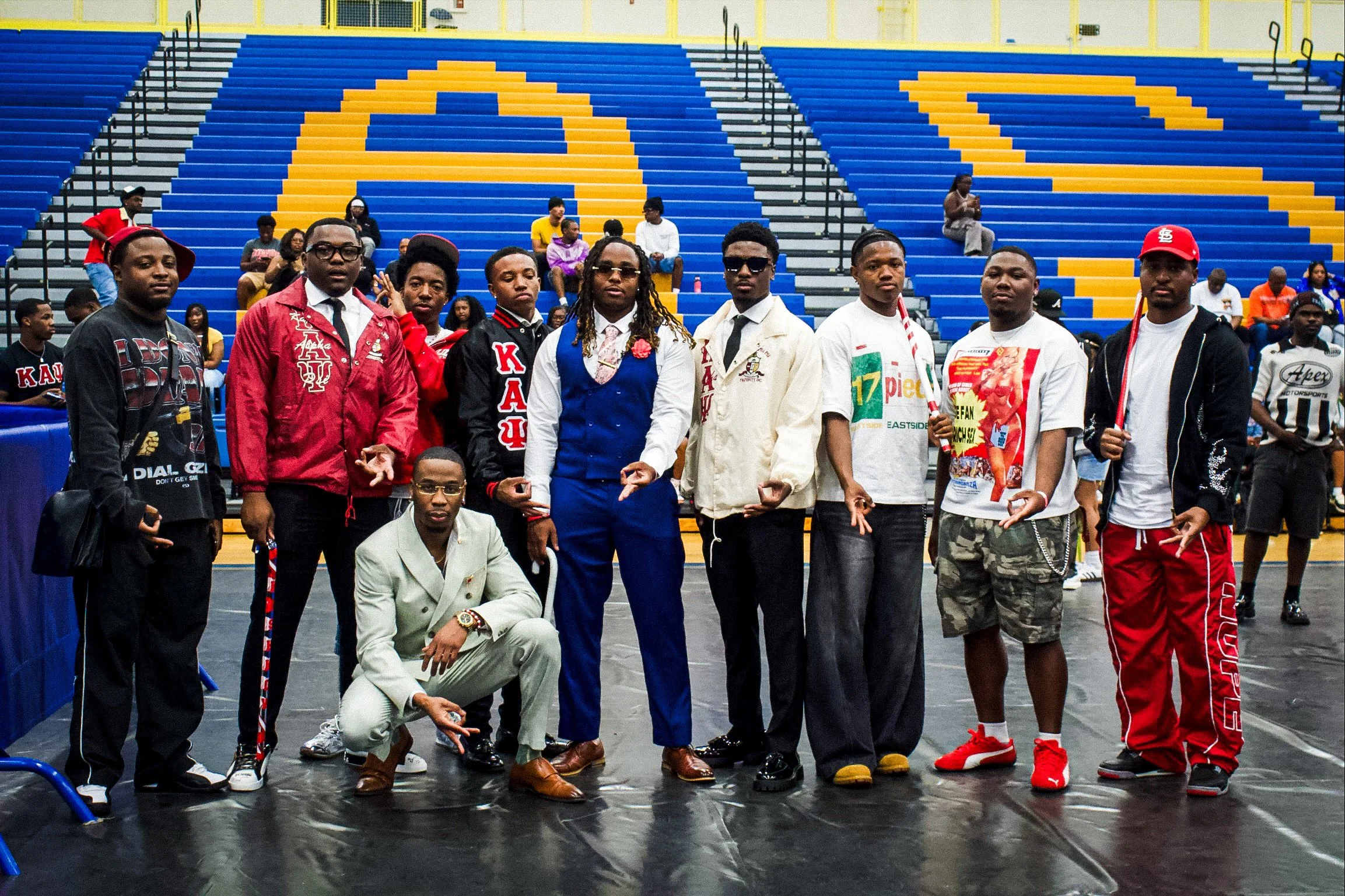 A group of young men in stylish clothing posing inside a gymnasium with blue and yellow bleachers and a few people sitting in the background.