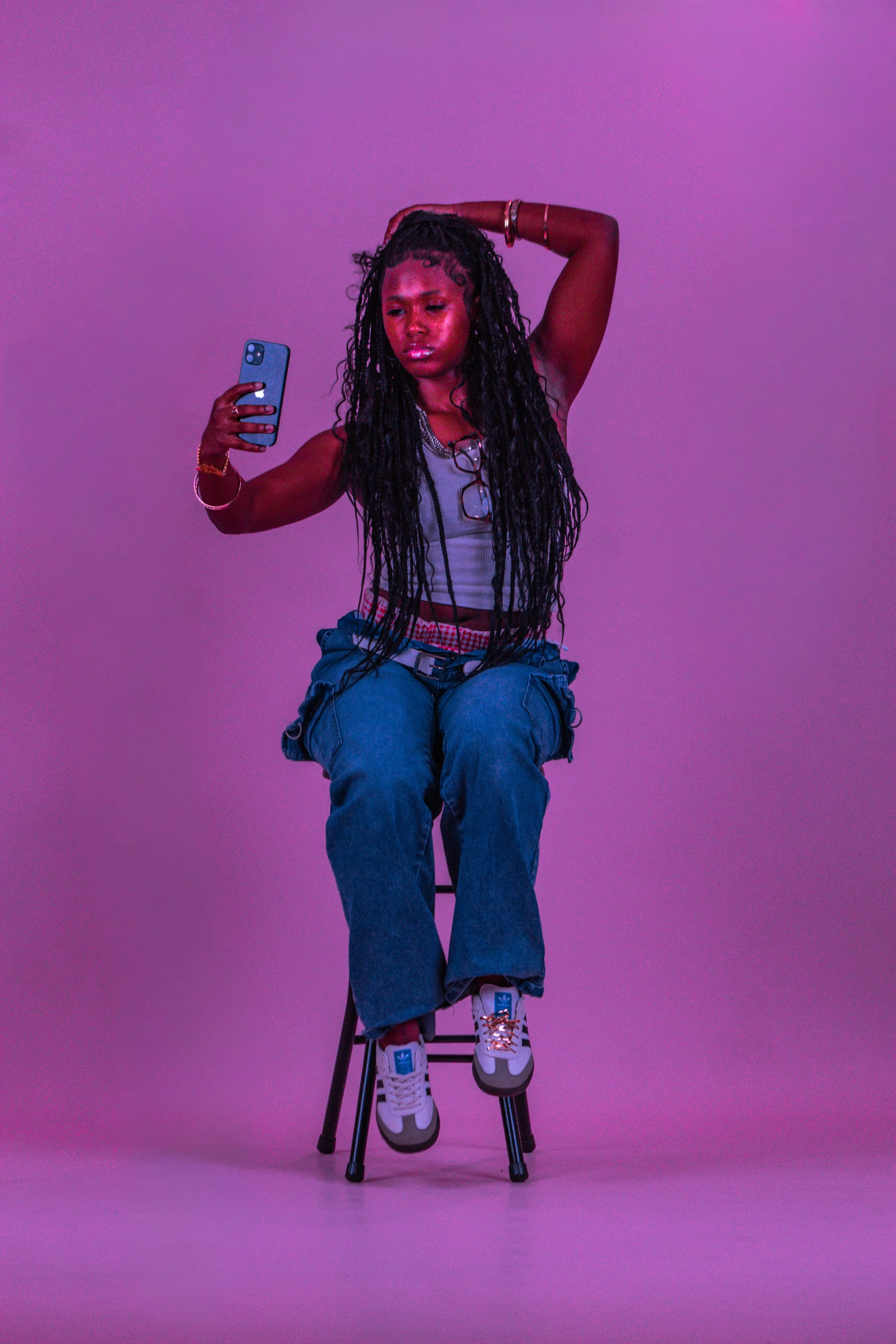 A young woman with long dreadlocks takes a selfie against a purple background. She is seated on a stool, wearing a white tank top, baggy blue jeans, and sneakers. She has accessories including bracelets, a necklace, and glasses hanging around her nec