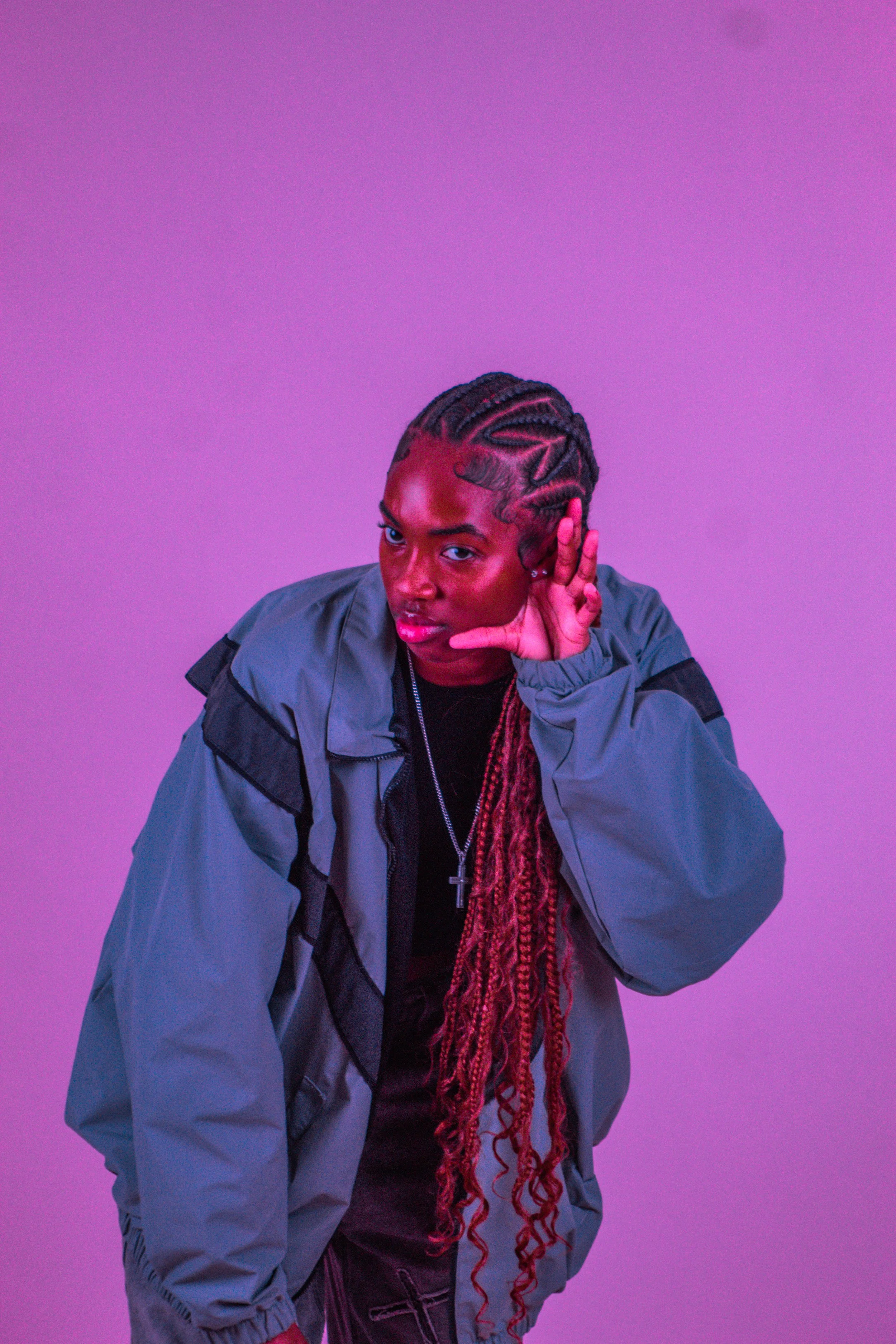 A person with braids and dreadlocks wearing a gray jacket, black shirt, and cross necklace, posing against a purple background.