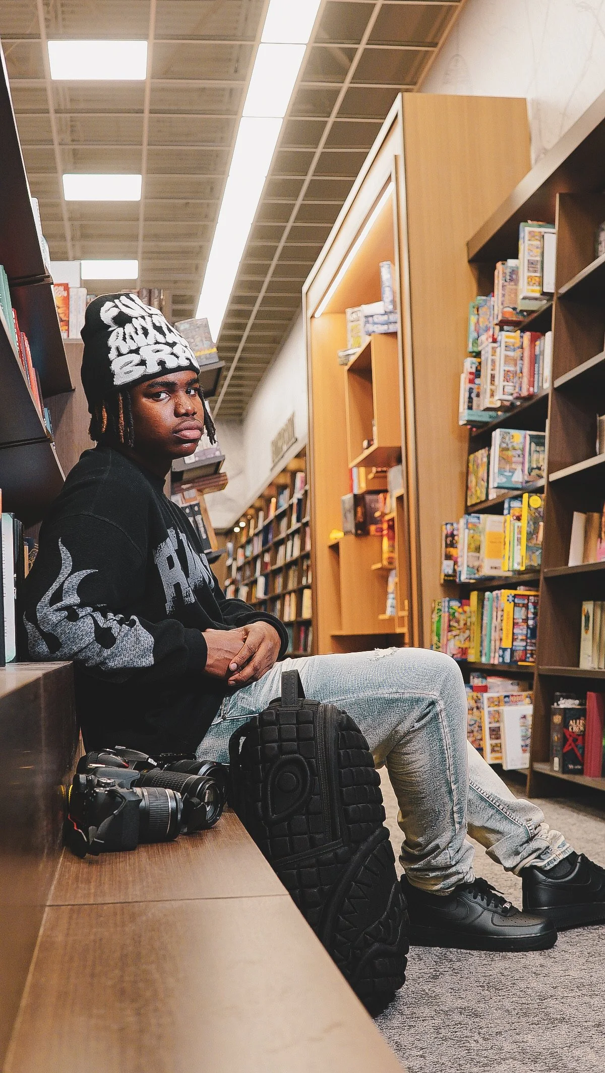 A young man sitting in a bookstore, wearing a black beanie, black hoodie, light blue jeans, and black sneakers, with camera gear including a camera and a vertical grip placed on the wooden bench next to him.