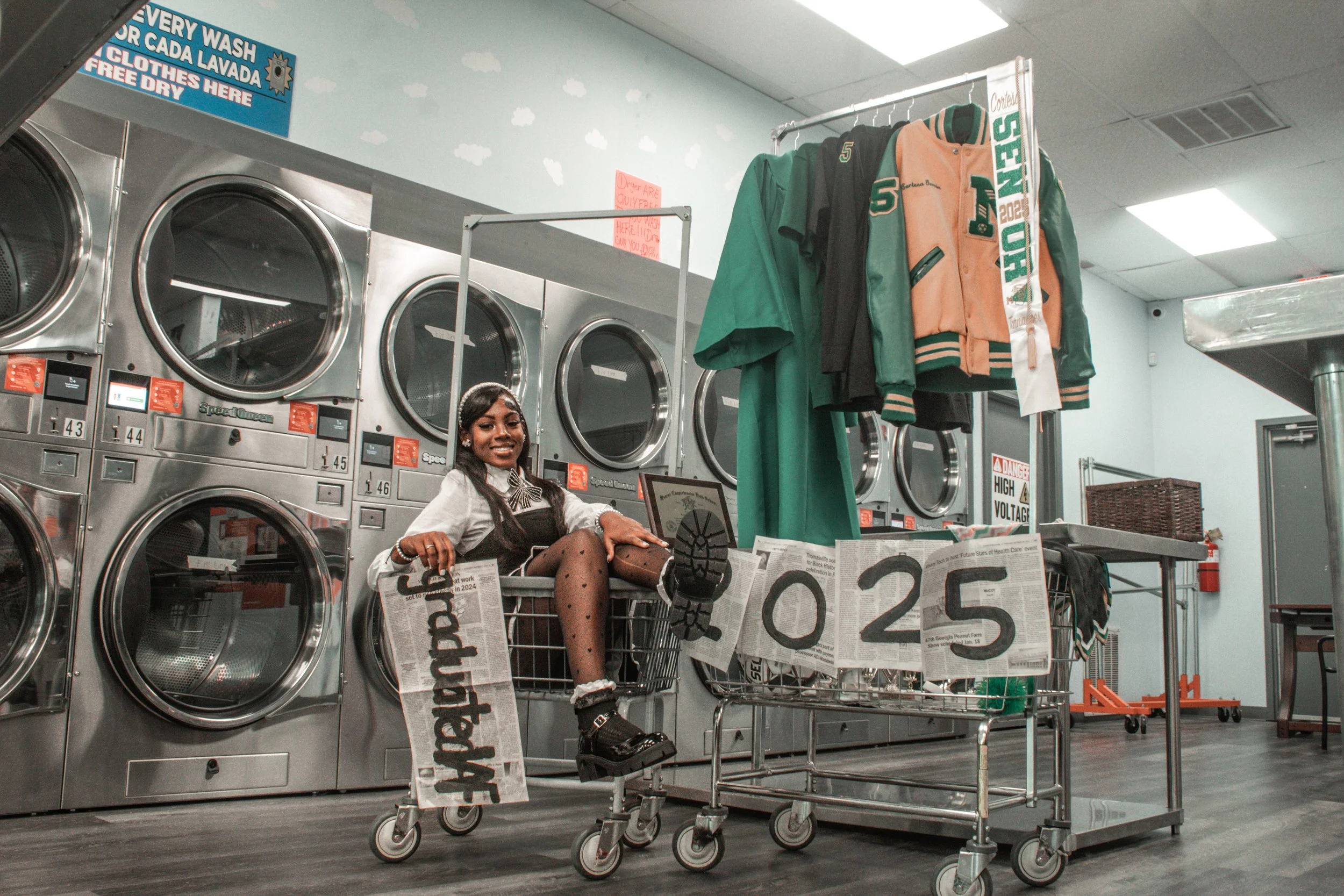 Inside a laundromat, a young woman is sitting in a laundry cart, smiling and looking at the camera. She's wearing a school uniform with a bow tie, tights with polka dots, and chunky boots. Surrounding her are laundry machines, signs, and hanging clot