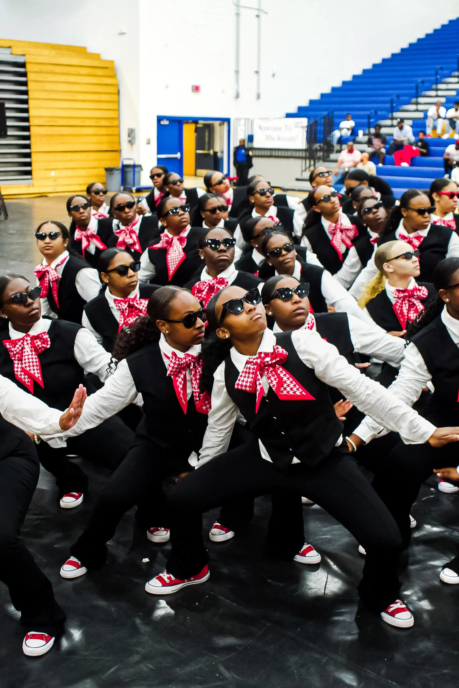 Group of women wearing black vests, white shirts, red and white checkered bandanas, and sunglasses, performing a choreographed dance in a gymnasium with blue bleachers and several spectators.