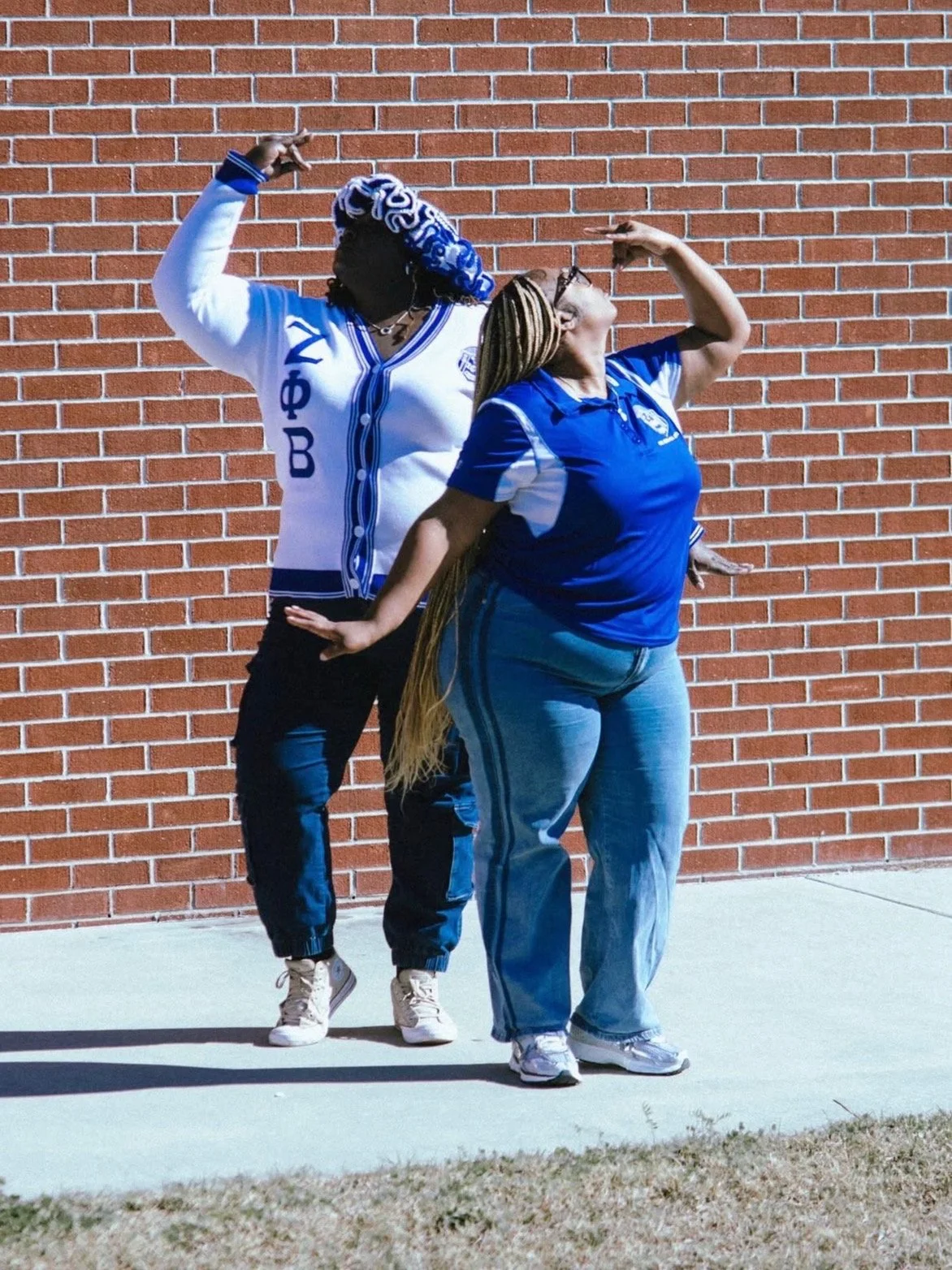 Two women are posing in front of a brick wall, standing back to back with their arms raised in a flexing pose, wearing blue and white school clothing.