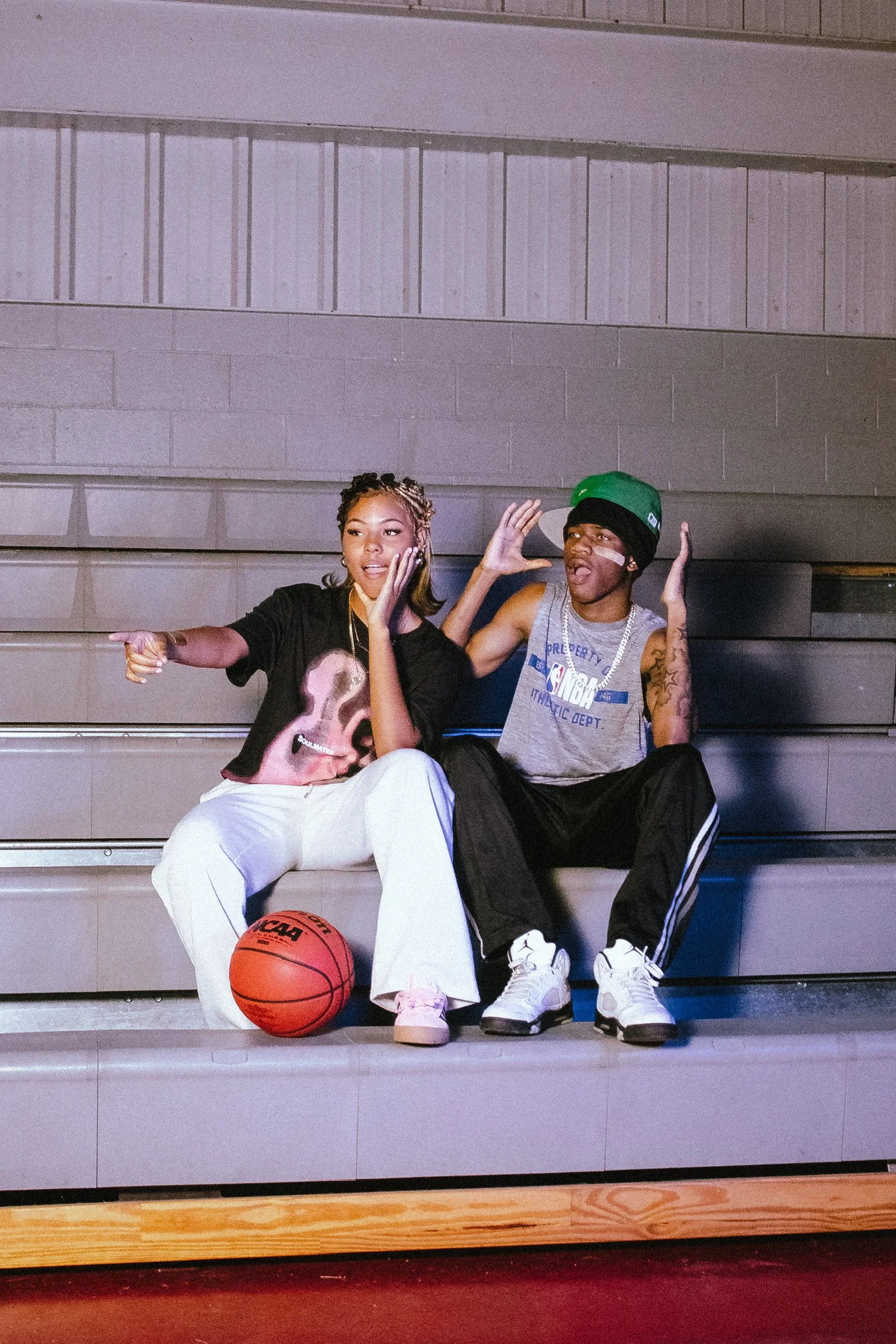 Two young people sitting on steps in a gymnasium or sports arena. The woman on the left has braided hair and is dressed in a black t-shirt and white pants, pointing and talking. The man on the right has a green cap, tattoos, a grey tank top, and blac