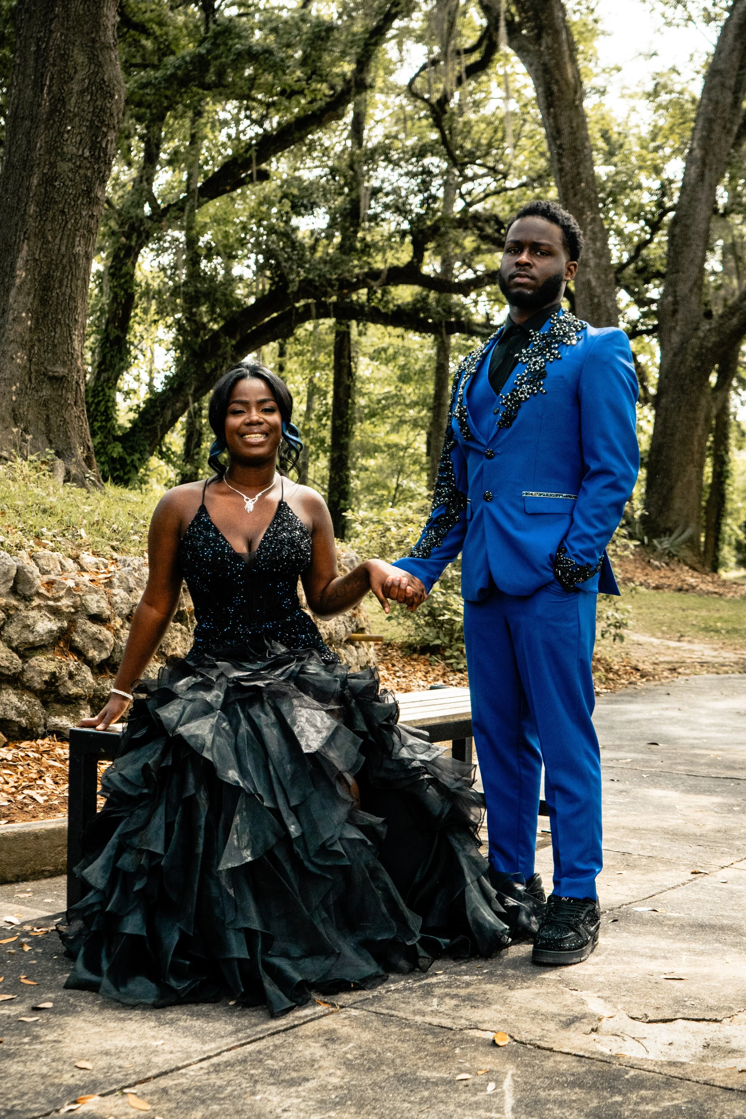A woman in a black sequined dress with a voluminous black skirt, sitting on a park bench, and a man in a bright blue suit with embellishments, standing next to her, holding her hand, with trees and greenery in the background.