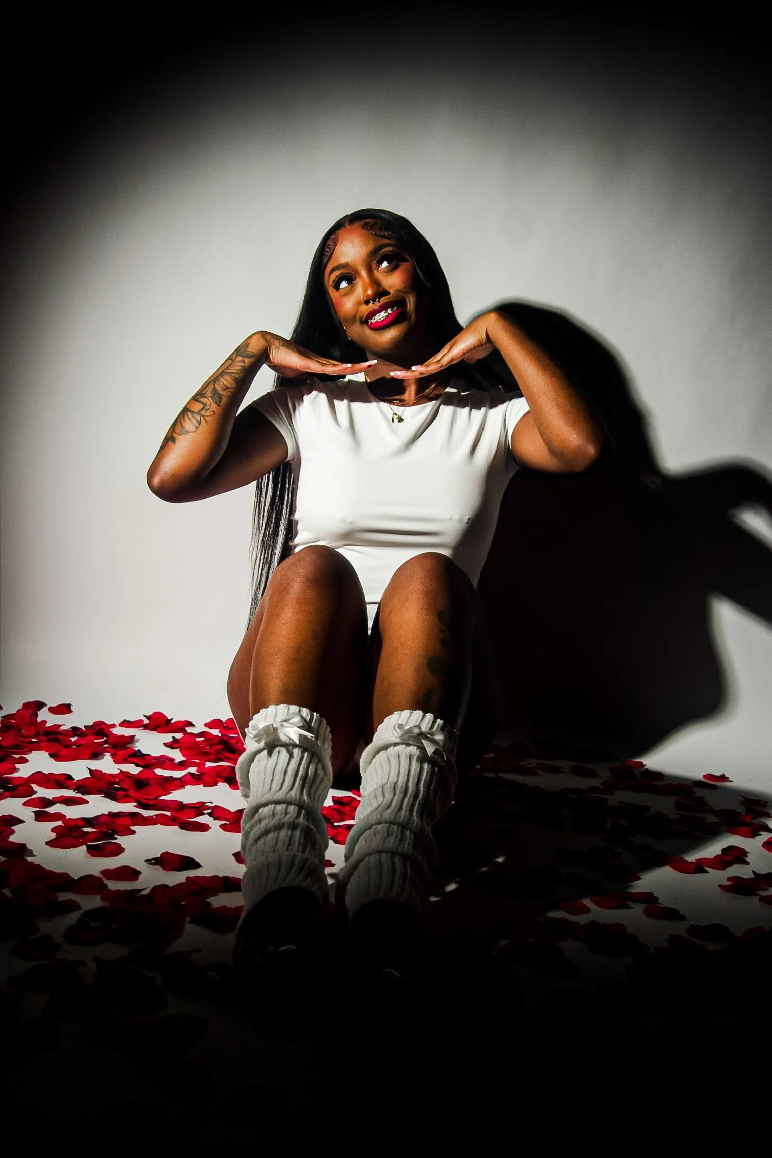 A woman with tattoos on her left arm and wearing a white shirt sits on the floor surrounded by red rose petals. She holds her hands near her chin, looking upward with a playful expression, and has a shadow cast on the plain white background. A person