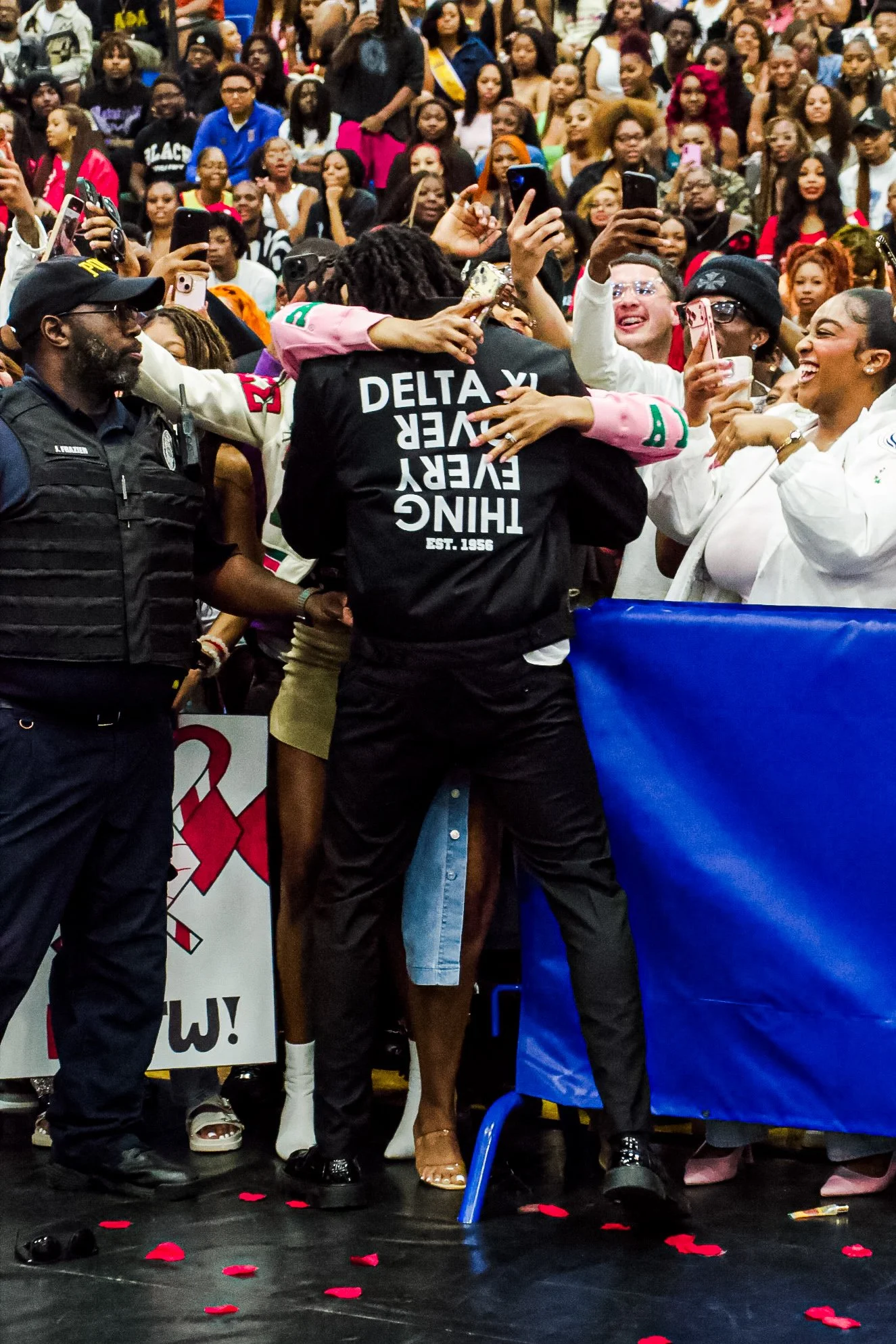 A large crowd of diverse people gathered indoors. Several individuals are taking selfies and photos, smiling and hugging. The scene appears celebratory, with rose petals scattered on the floor.
