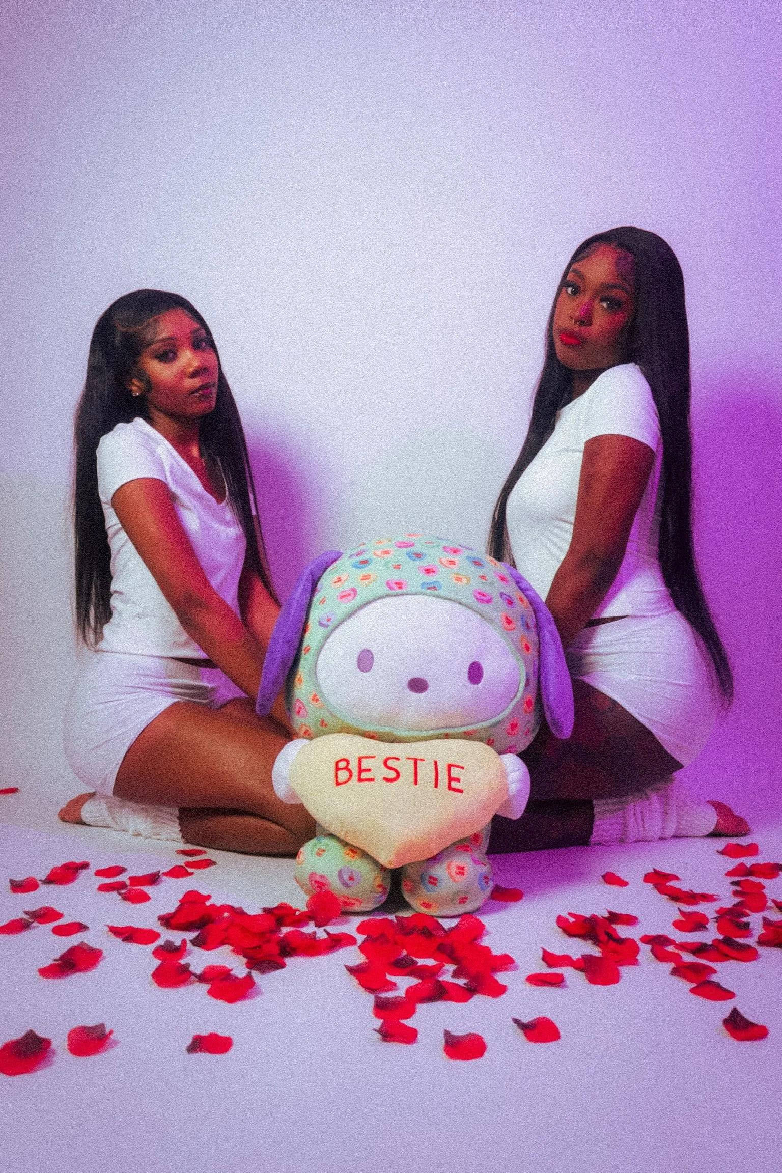 Two women in white outfits kneeling on the floor surrounded by red rose petals, holding a plush toy of a cute animal with a sign that reads 'BESTIE'.
