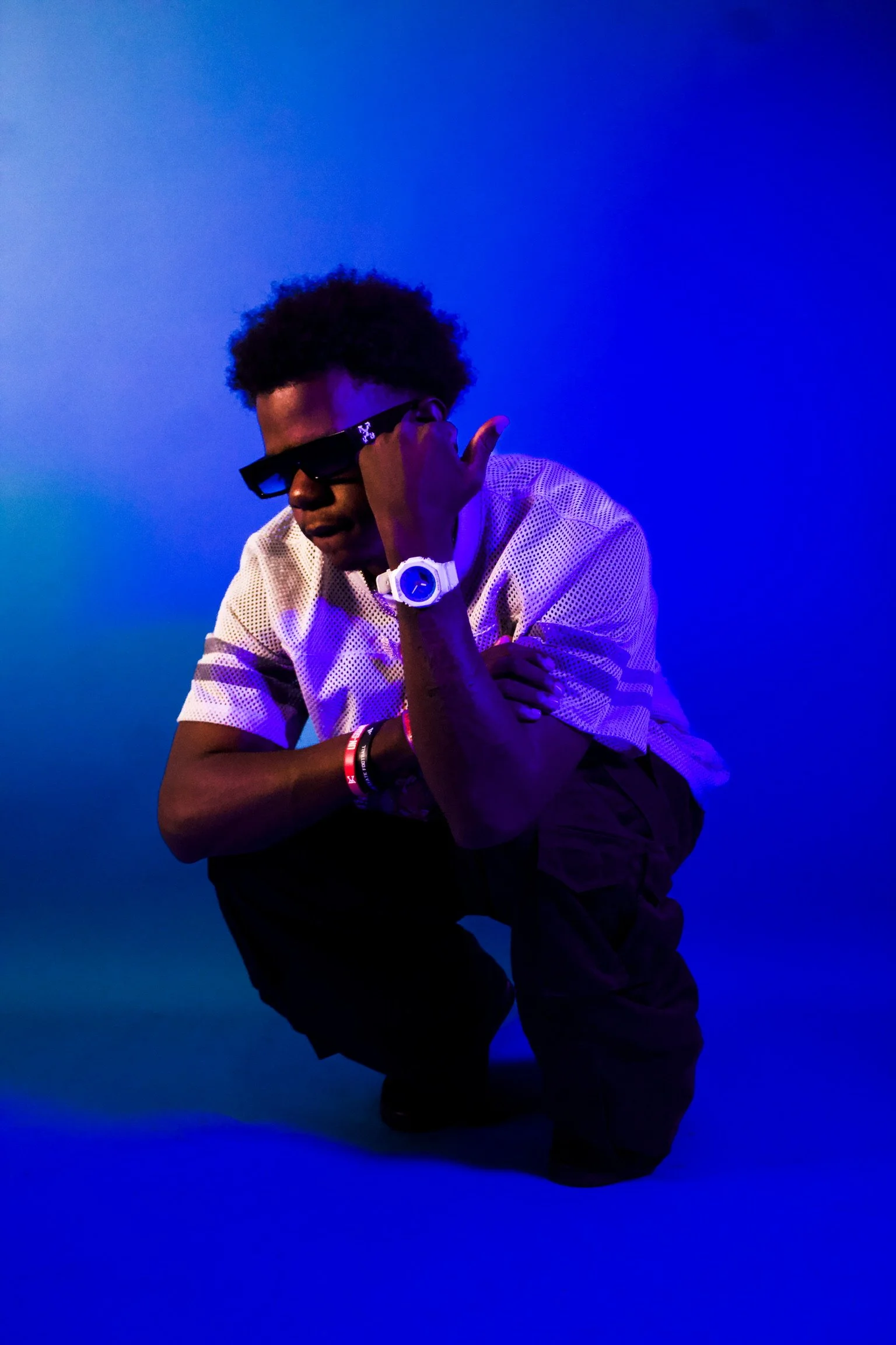 A person with dark curly hair wearing dark sunglasses, a white short-sleeve shirt, and dark pants, is crouching on one knee against a gradient blue background, with one hand touching their face and the other arm crossed over their chest.