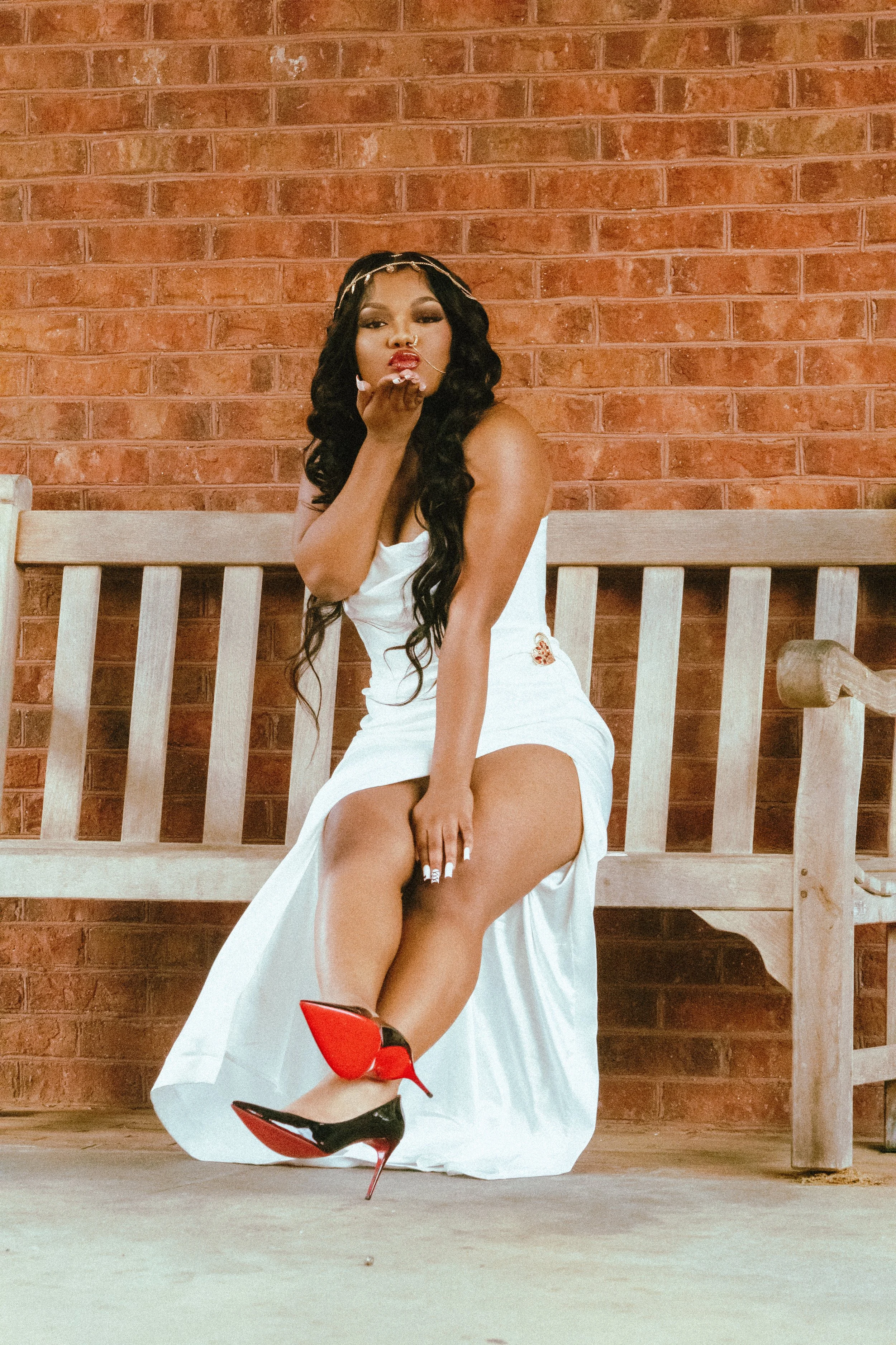 A woman with long dark curly hair, wearing red and black high heels and a white dress, sitting on a wooden bench with a brick wall background, blowing a kiss toward the camera.