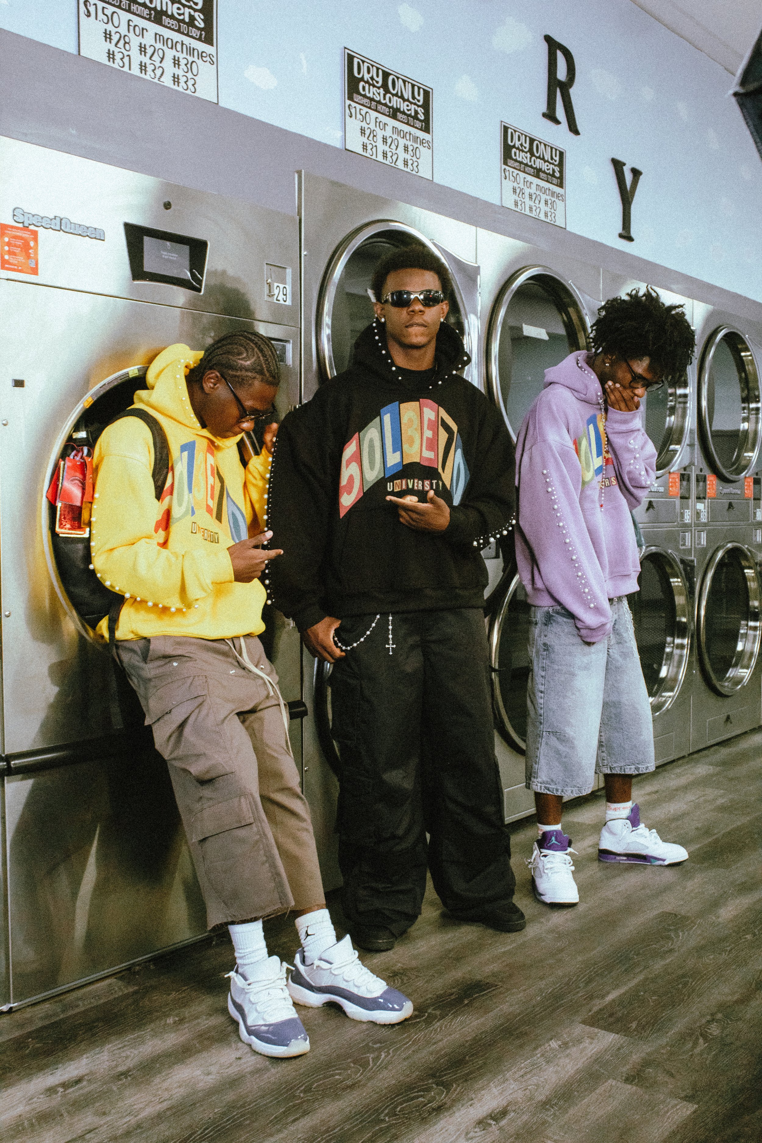 Three young men standing in a laundromat, wearing colorful hoodies and sneakers, with laundry machines behind them.